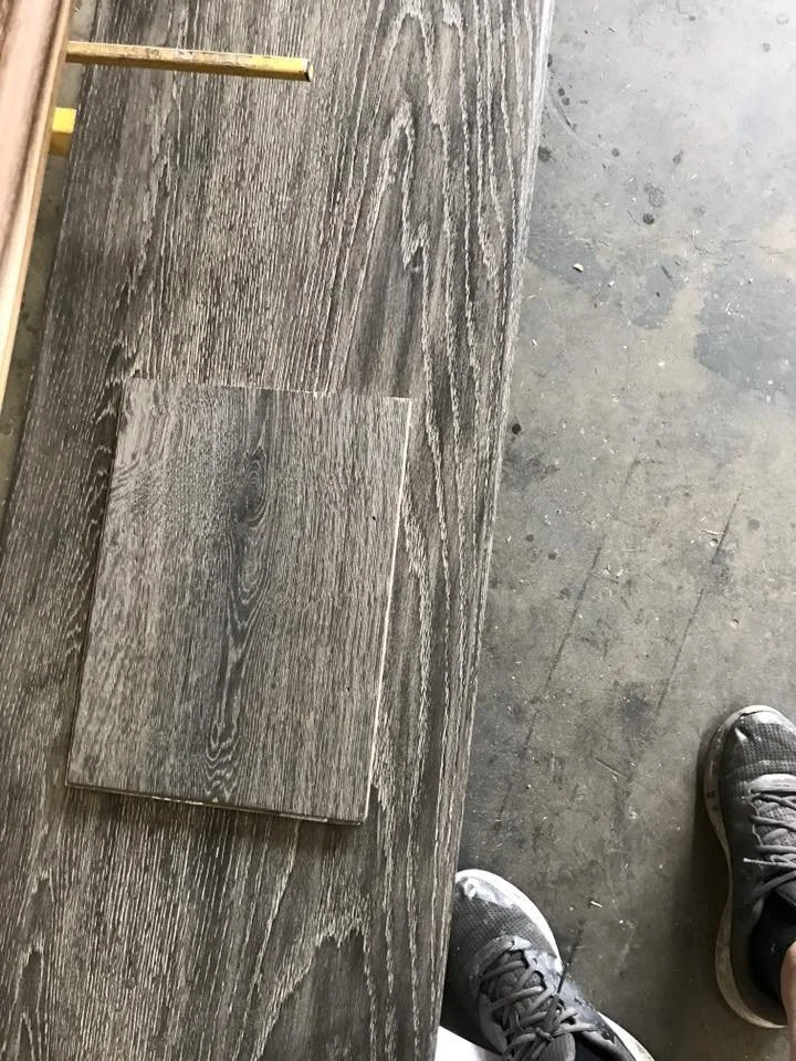 Interlocking gray wood-look vinyl planks on the floor near a concrete floor with a pair of gray sneakers visible at the bottom right corner.