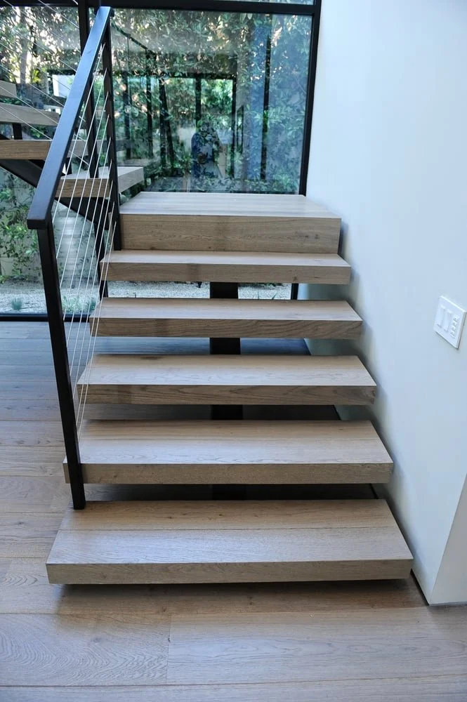 Modern staircase with wooden steps, metal railing with horizontal cables, adjacent glass window showing greenery outside, and a white wall with electrical outlet.