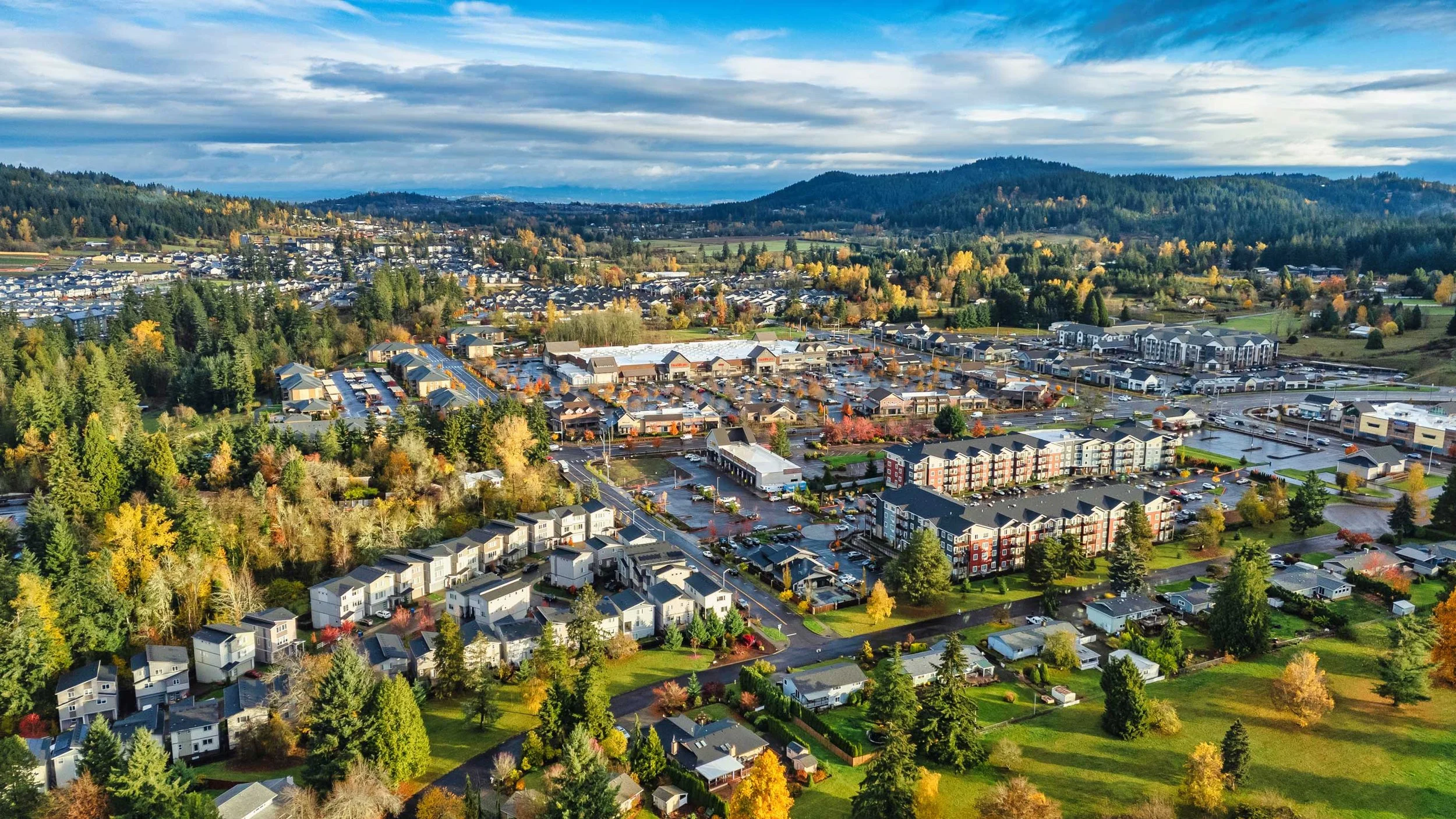 Aerial view of a town with a mix of residential homes, shopping centers, and parking lots amidst a landscape of green trees and rolling hills under a partly cloudy sky.