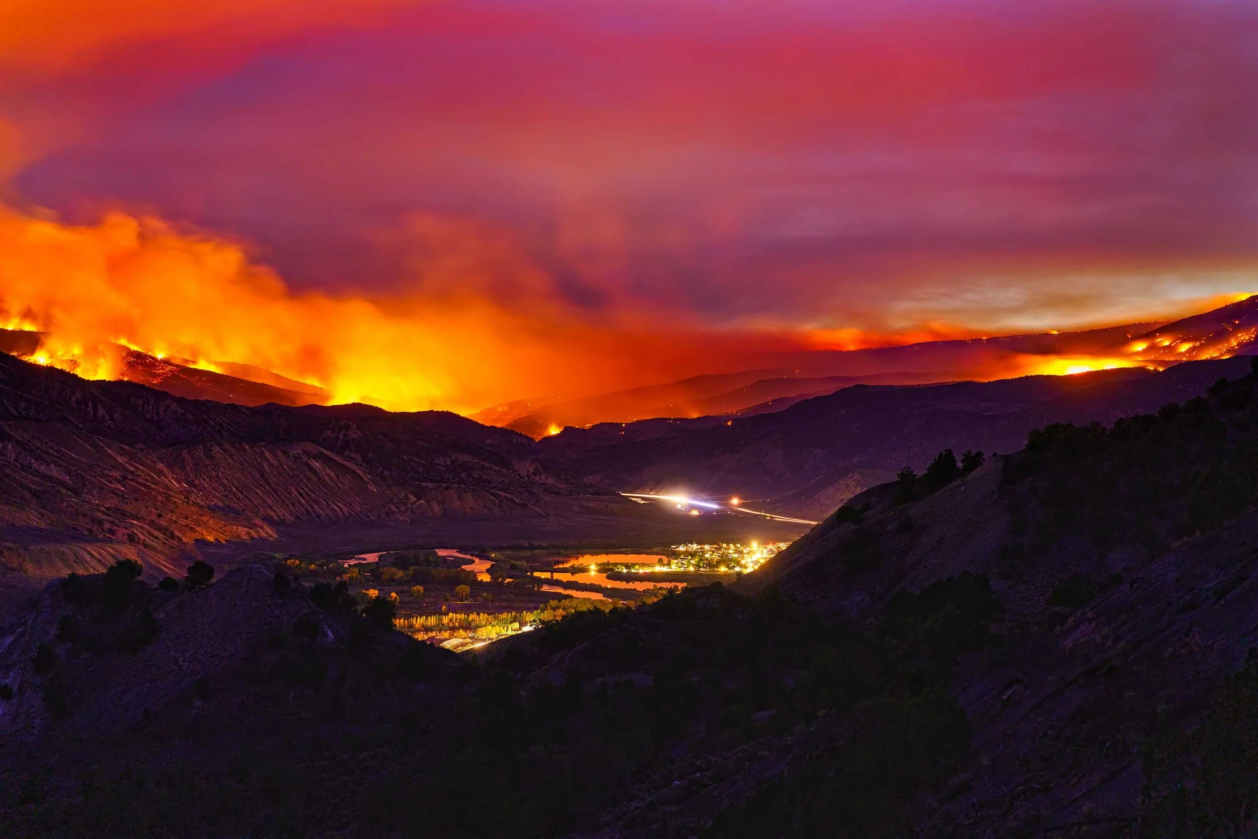 Nighttime landscape of a wildfire burning on a hillside with a purple and orange sky, illuminating smoke and flames, with a river and small town in the valley below.