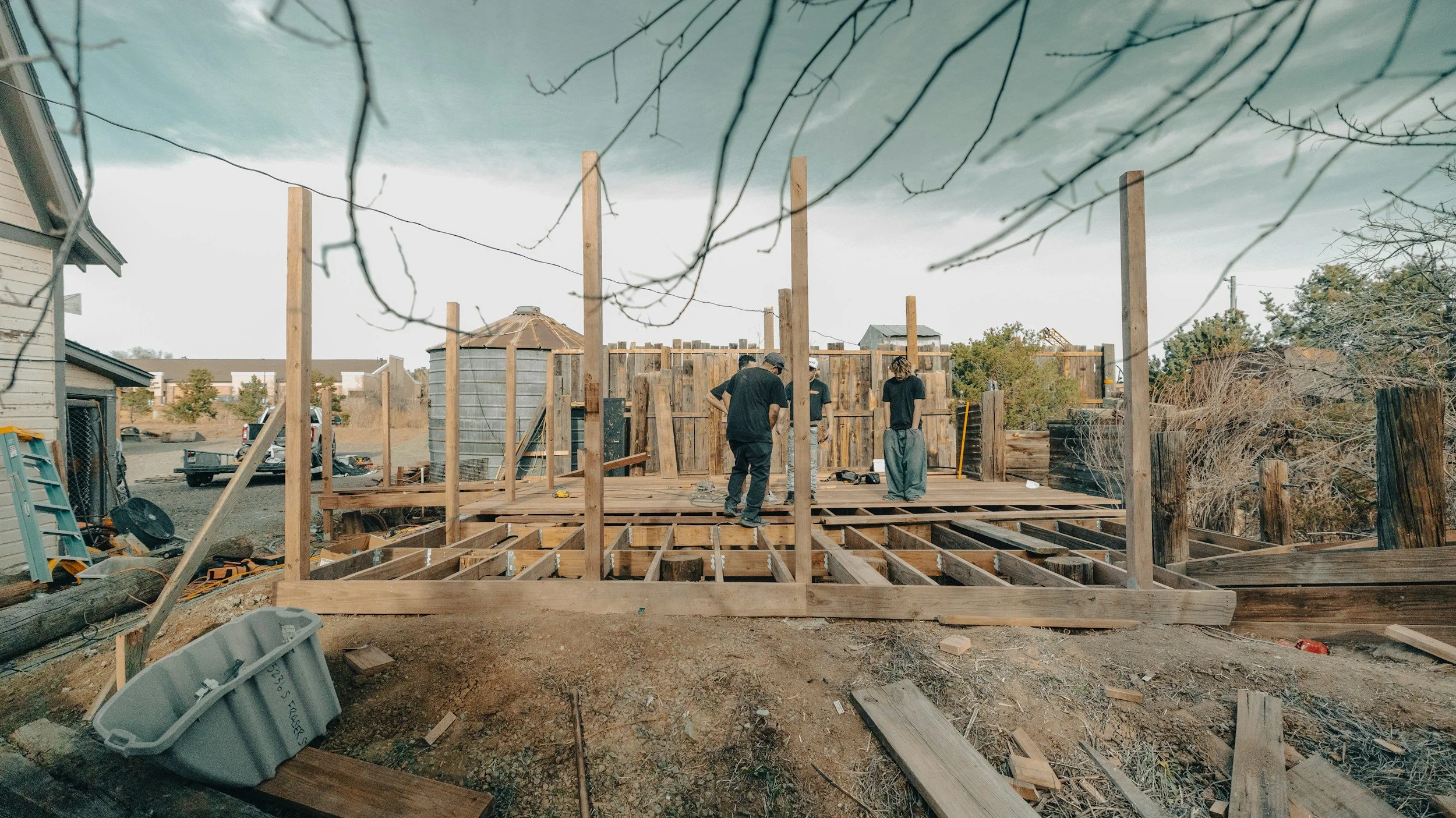 Construction workers building a wooden structure on a residential lot, with a cloudy sky above and various building materials scattered around.