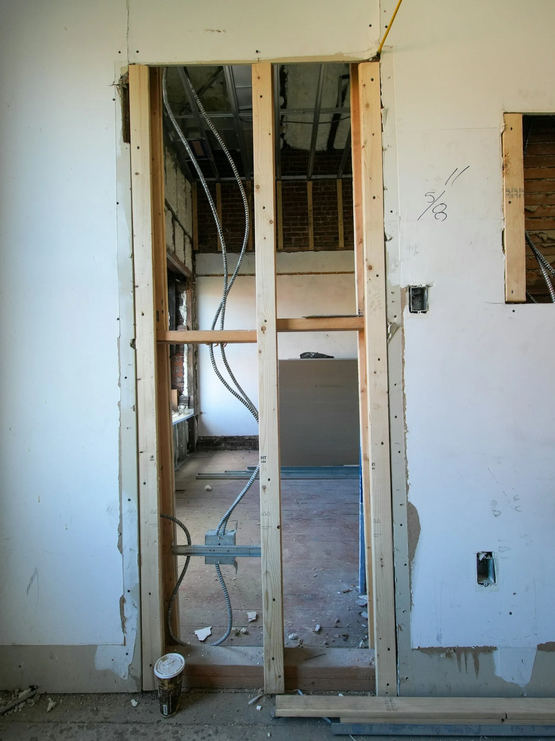 View of a room under construction with exposed wooden framing for a doorway and electrical wiring visible. The surrounding walls are partially finished with some markings, and a coffee cup is on the floor.