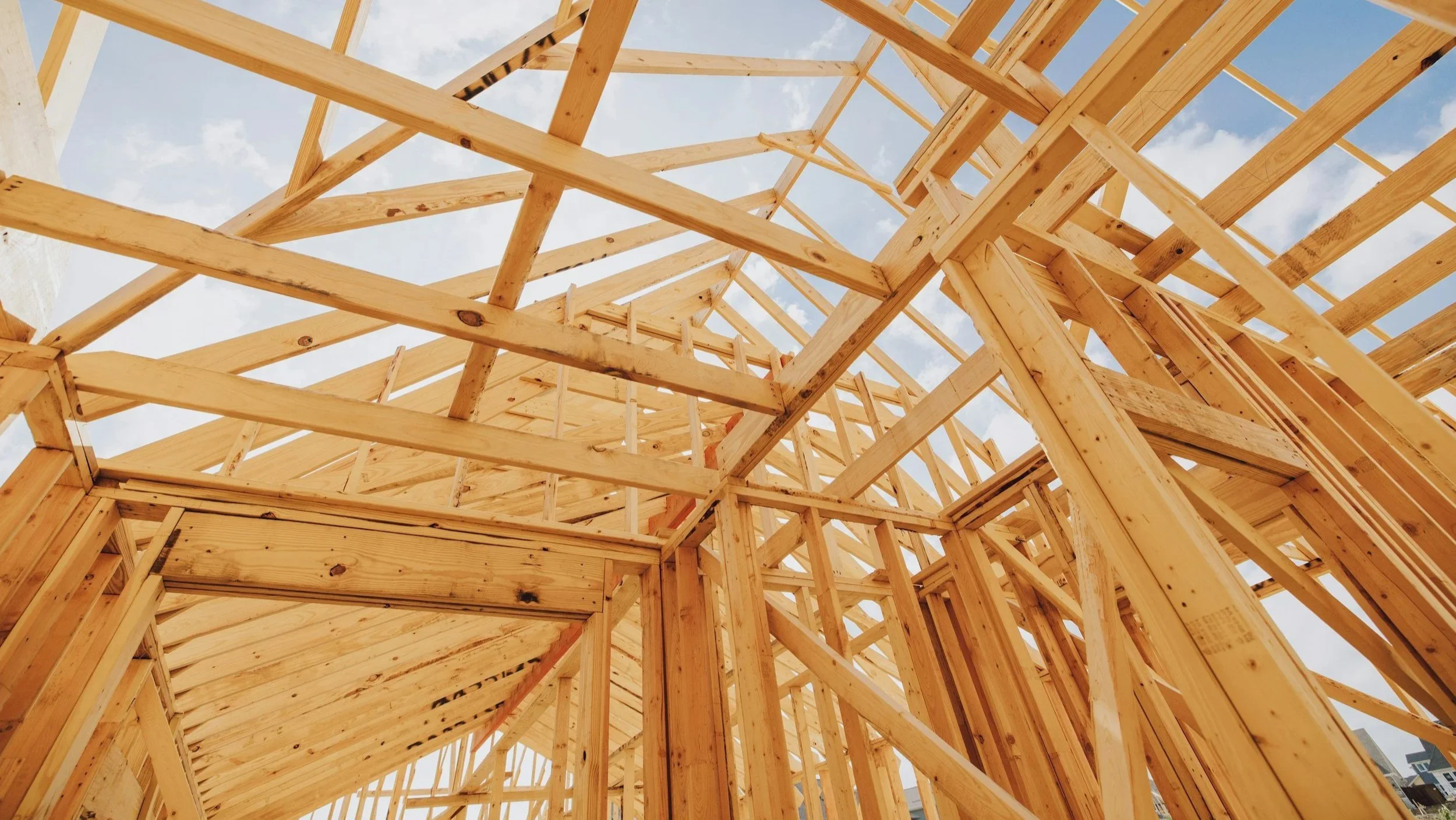 Wooden framing of building under construction, viewed from below with a blue sky and clouds in the background.