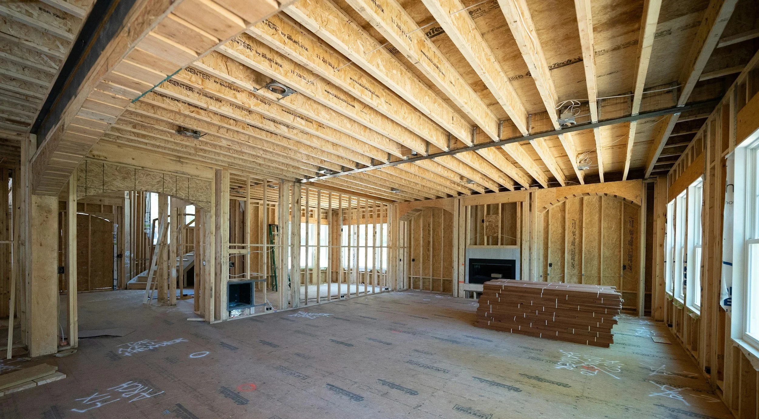 Interior of a house under construction showing exposed wooden framing, including ceiling joists and wall studs, with some construction markings on the floor and stacked construction materials.