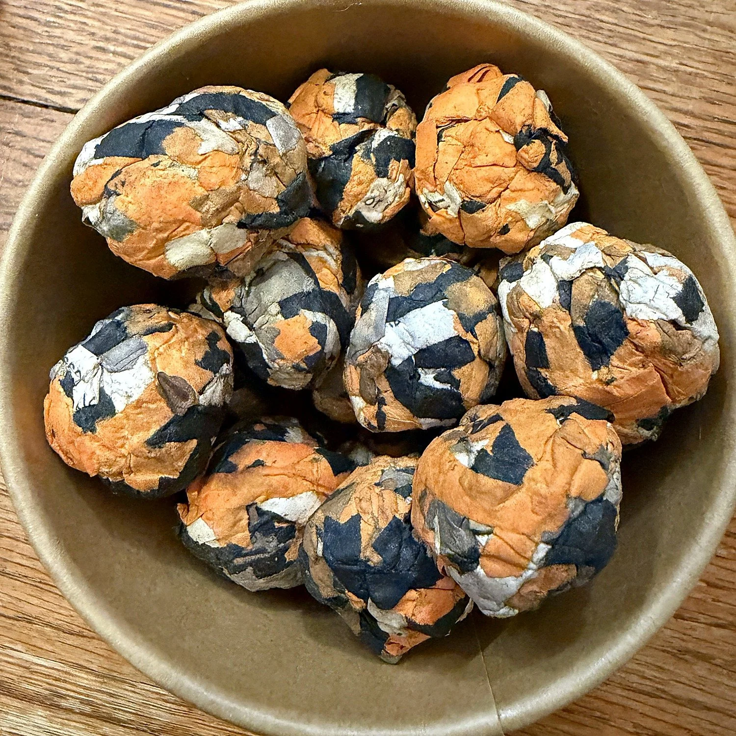 Orange and black milkweed seed balls inside a kraft gift pot, seen from above.