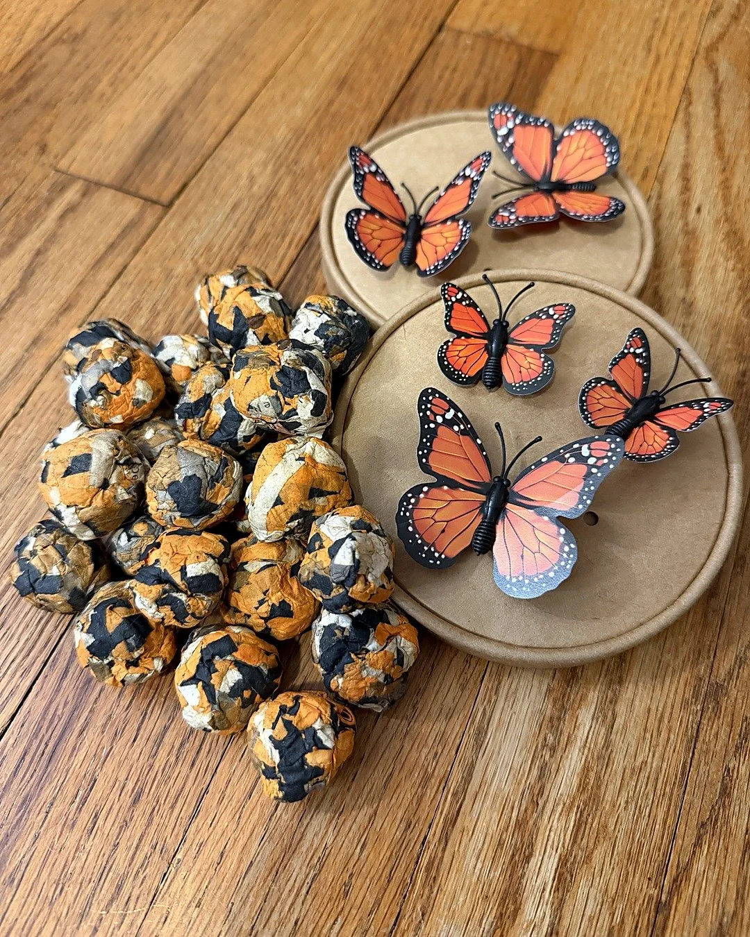 Orange and black recycled-paper milkweed seed balls piled beside two round kraft lids topped with 3D monarch butterflies.