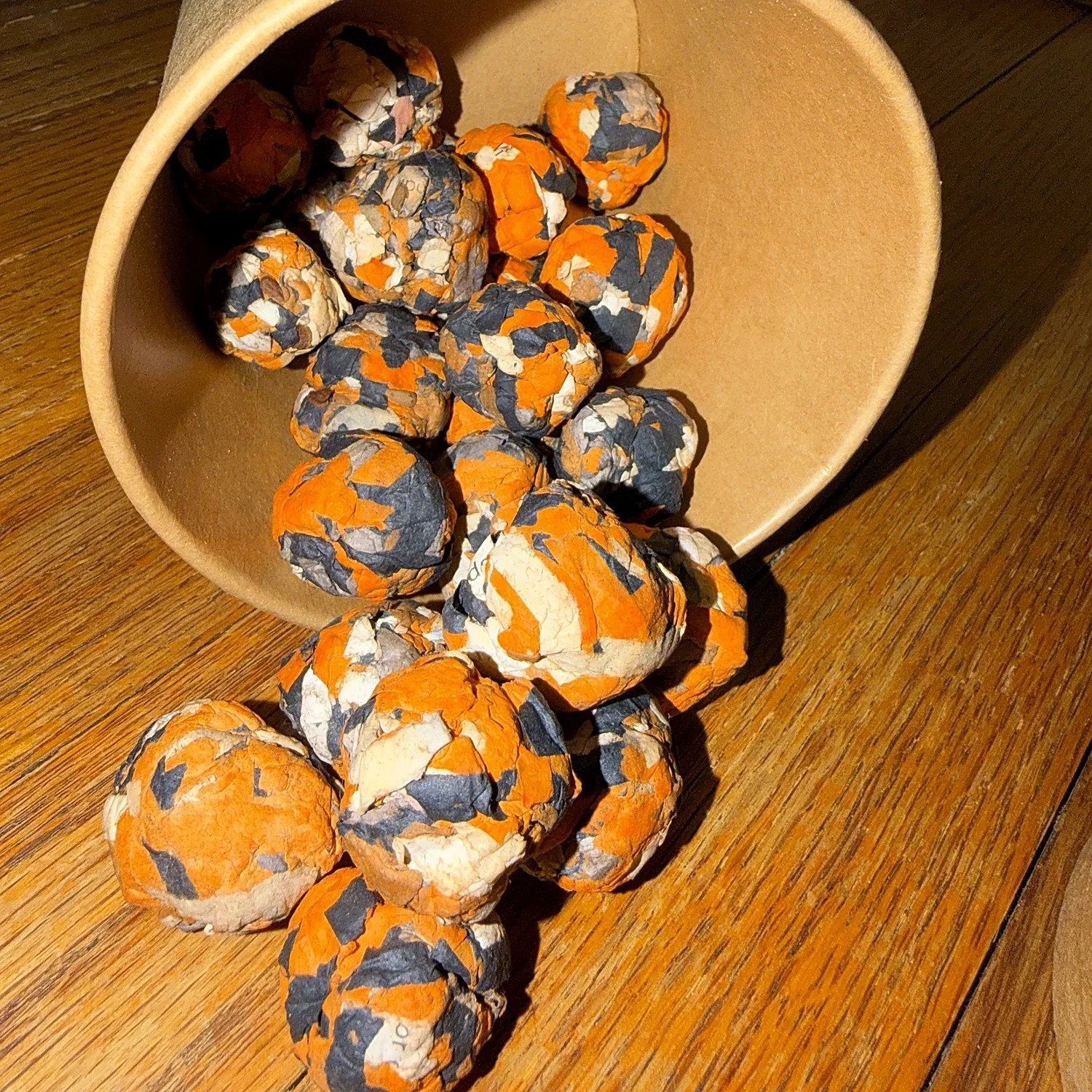 Orange and black milkweed seed balls spilling from a kraft gift pot beside its monarch lid.