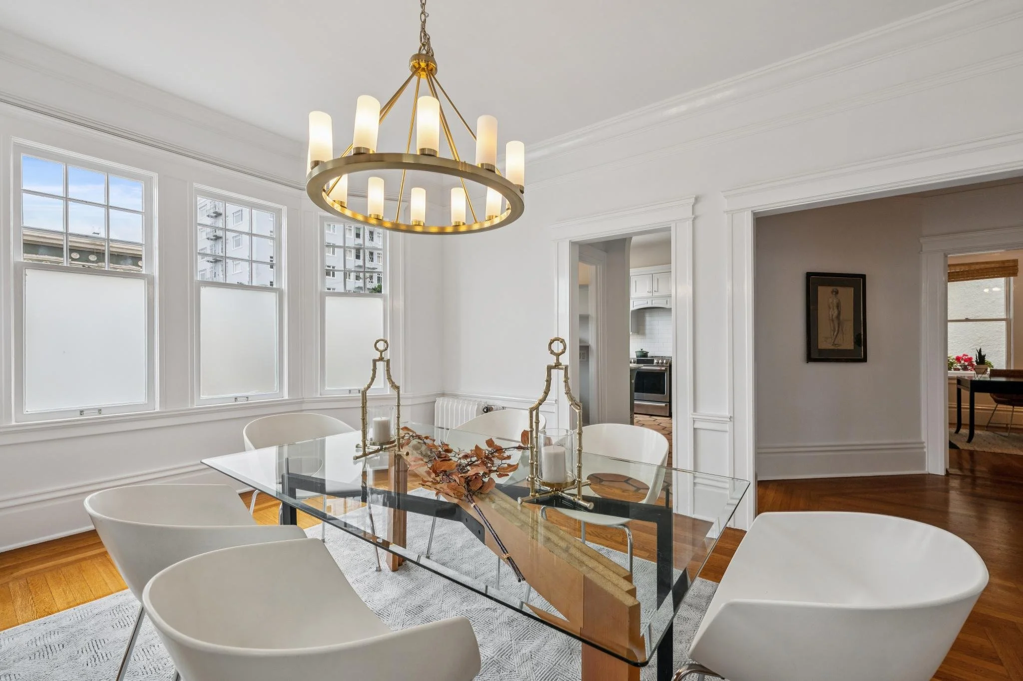 Modern dining room with a glass-top table, white chairs, a gold chandelier, and large windows with white blinds, in a bright, white interior.