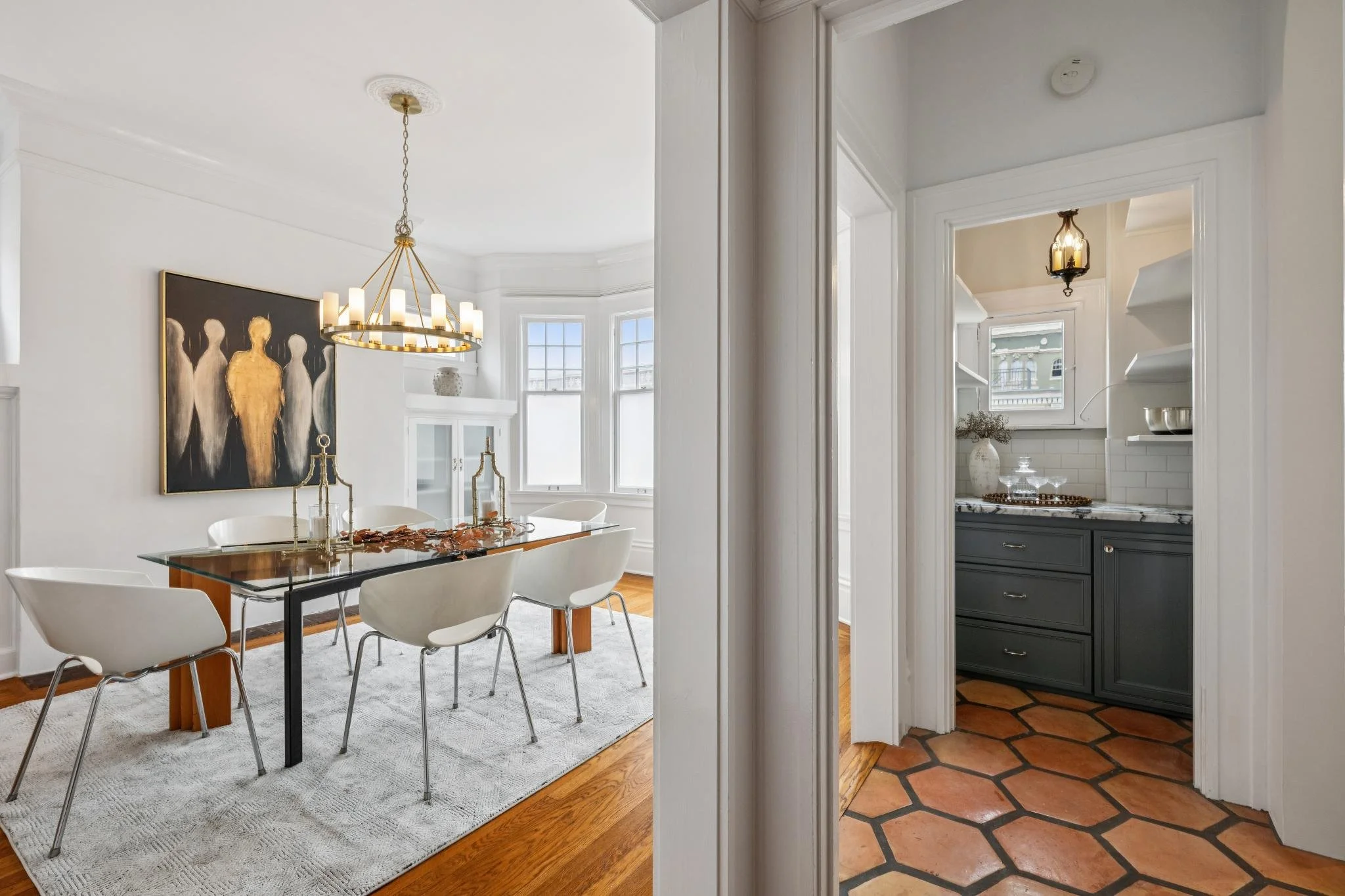 Dining room with a glass table, white chairs, a chandelier, and a large abstract art piece on the wall. Kitchen with black cabinets and tiled floor visible through doorway.