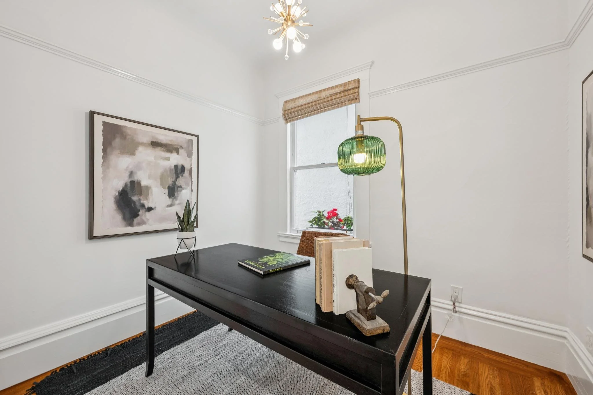 Interior corner of a room with a black table, abstract art, a potted plant, books, a green glass lamp, and decorative items, white walls, a window with bamboo shade, and a modern chandelier.