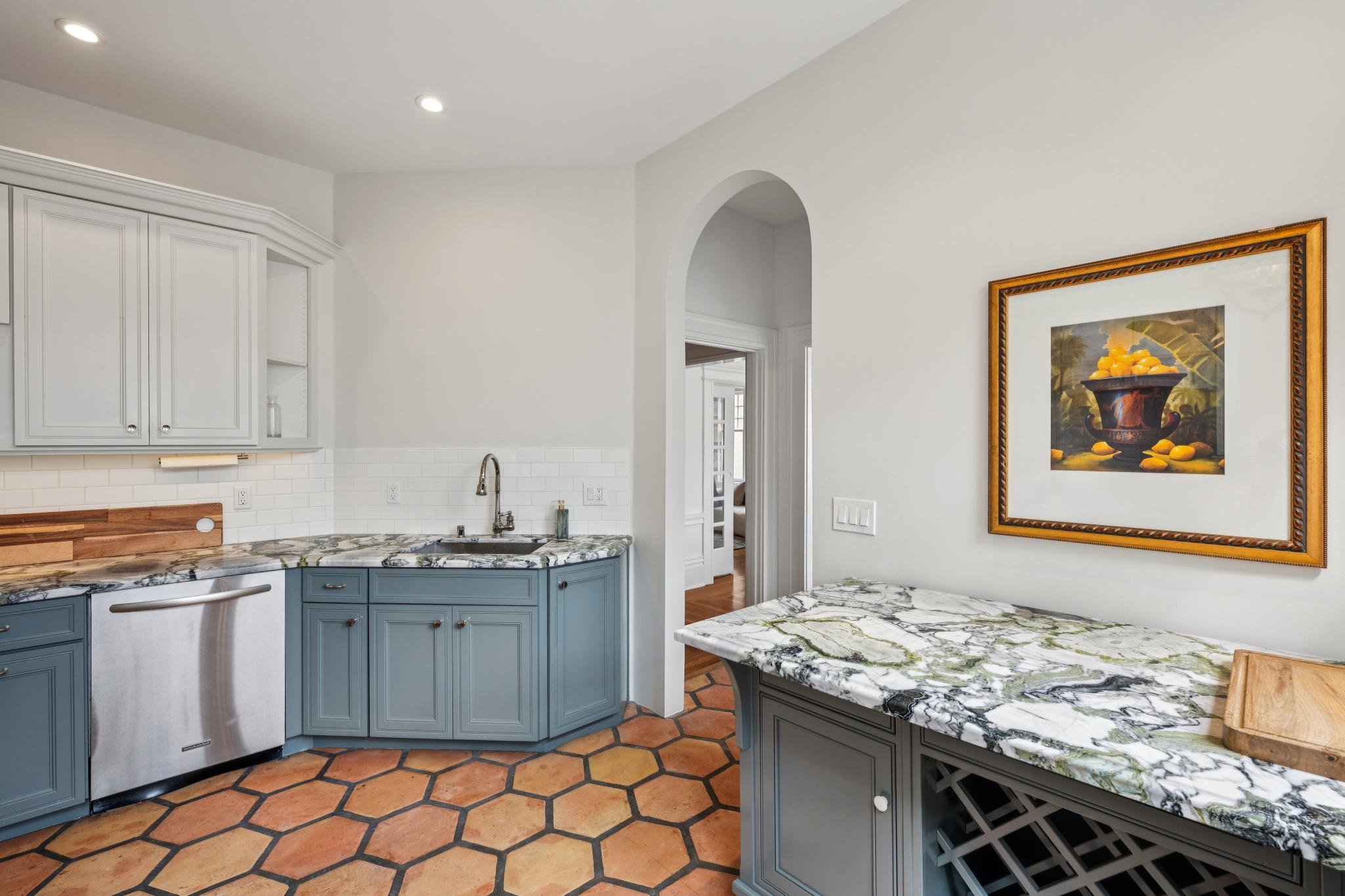 A kitchen with light gray cabinets, a marble countertop, a stainless steel dishwasher, and terracotta tile flooring. There is a framed painting of a black bowl with yellow fruit on the wall, and an arched doorway leading to another room.