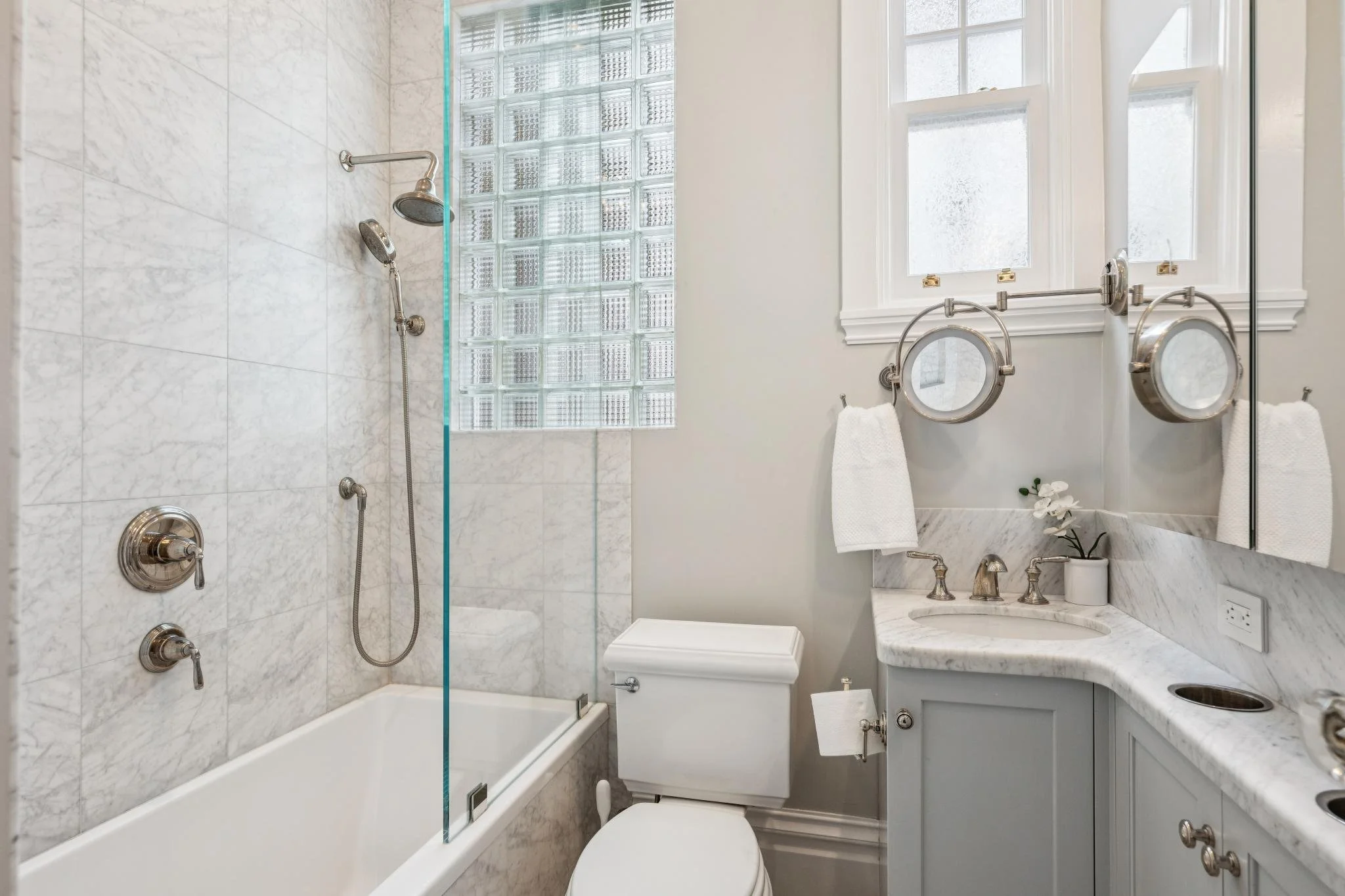 A modern bathroom with white marble walls and a bathtub with a glass segment. There is a large mirror above a white sink with a marble countertop, and a small potted plant on the counter. A toilet is next to the vanity, and two round magnifying mirro