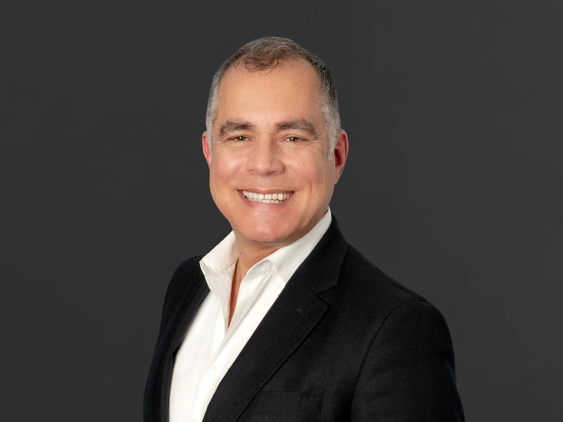 Portrait of a smiling middle-aged man with short hair, wearing a black suit and white shirt, against a dark gray background.