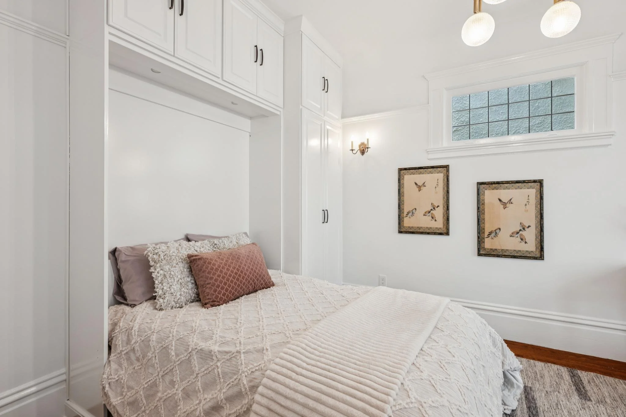 A bedroom with white walls, a bed with textured white bedding, and pillows including a pink patterned and a white fluffy one. There are framed bird artwork on the wall, a wall-mounted light fixture, a window with textured glass, and ceiling lights.