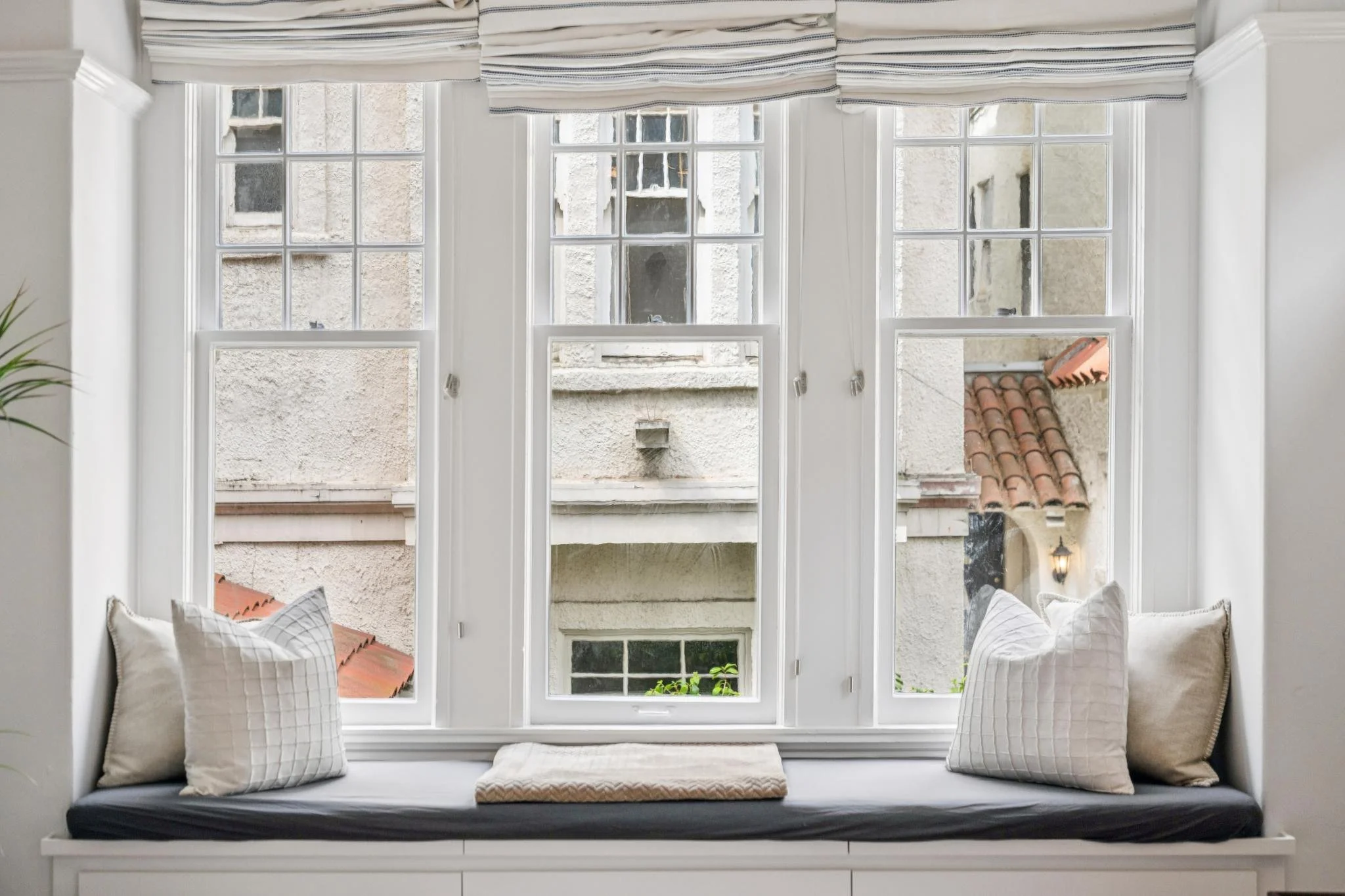 A cozy window seat with cushions and pillows, overlooking a view of neighboring buildings and rooftops through multiple-paned windows.