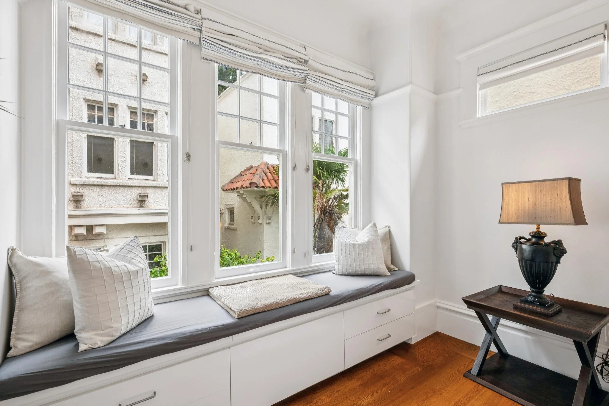 A cozy window seat decorated with pillows and a beige cushion, with a view of neighboring buildings and greenery outside the large multi-pane windows in a bright white room.