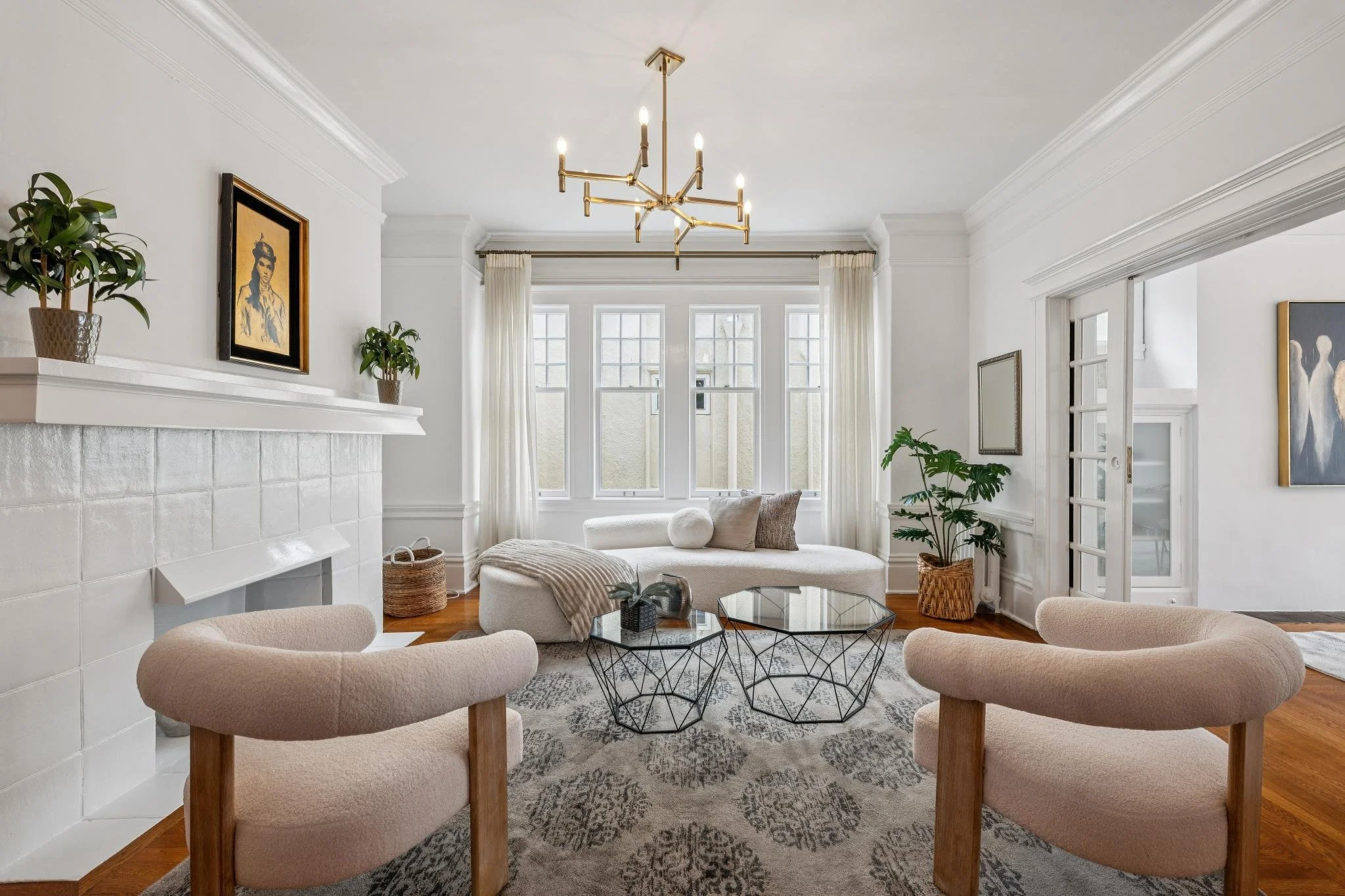 Bright and airy living room with white walls, large windows, modern chandelier, beige armchairs, round coffee tables, artwork on walls, plants, and neutral decor.