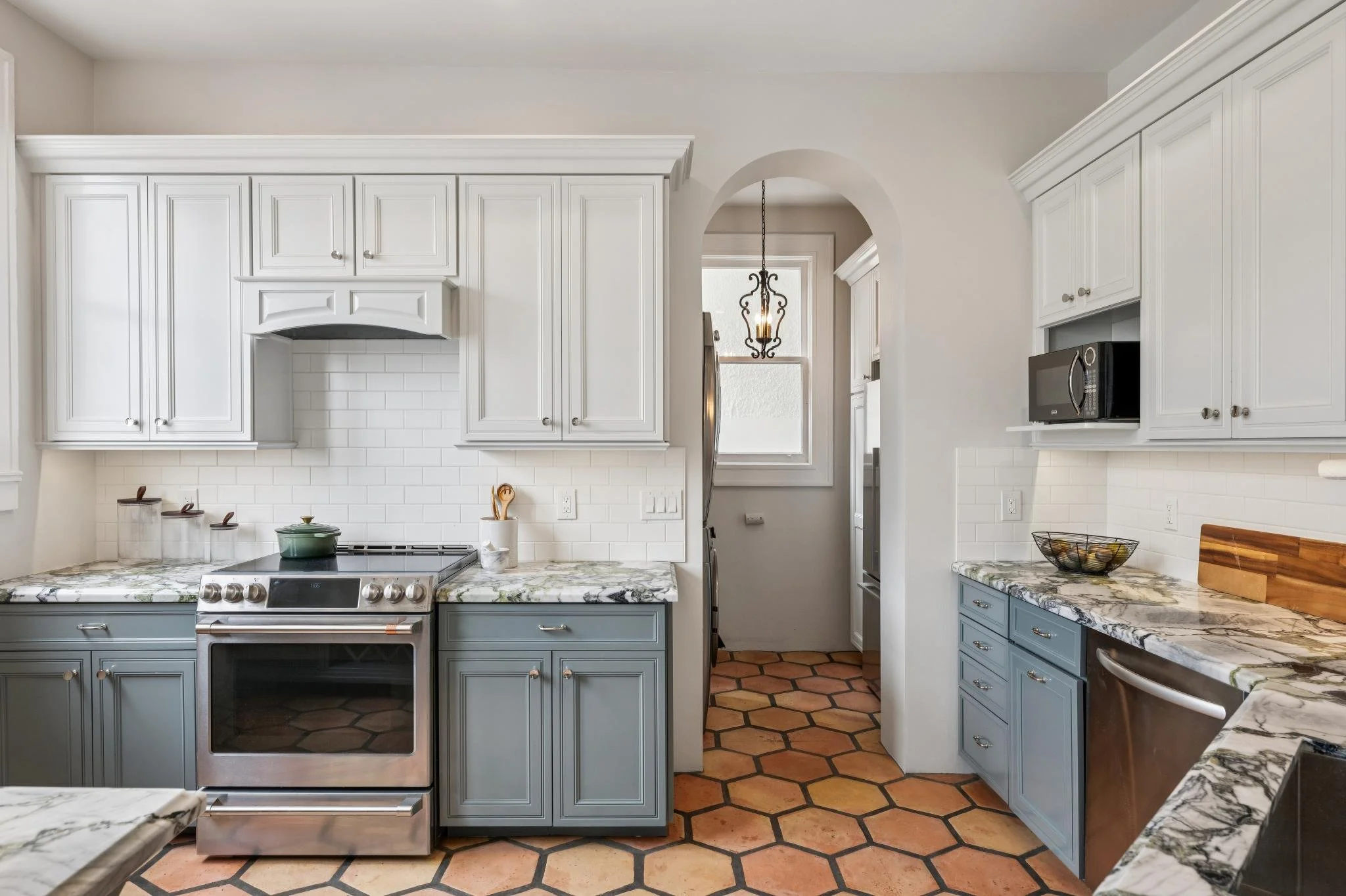 Kitchen with white upper cabinets, blue lower cabinets, marble countertops, a stove, a microwave, and terracotta tiled floor.