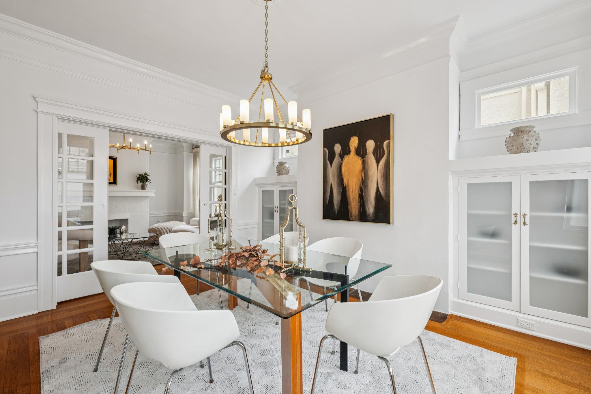 A dining room with a glass-top table and six white chairs. A gold chandelier hangs above the table. The room has white walls, built-in cabinets, and artwork on the wall. There are windows letting in natural light.