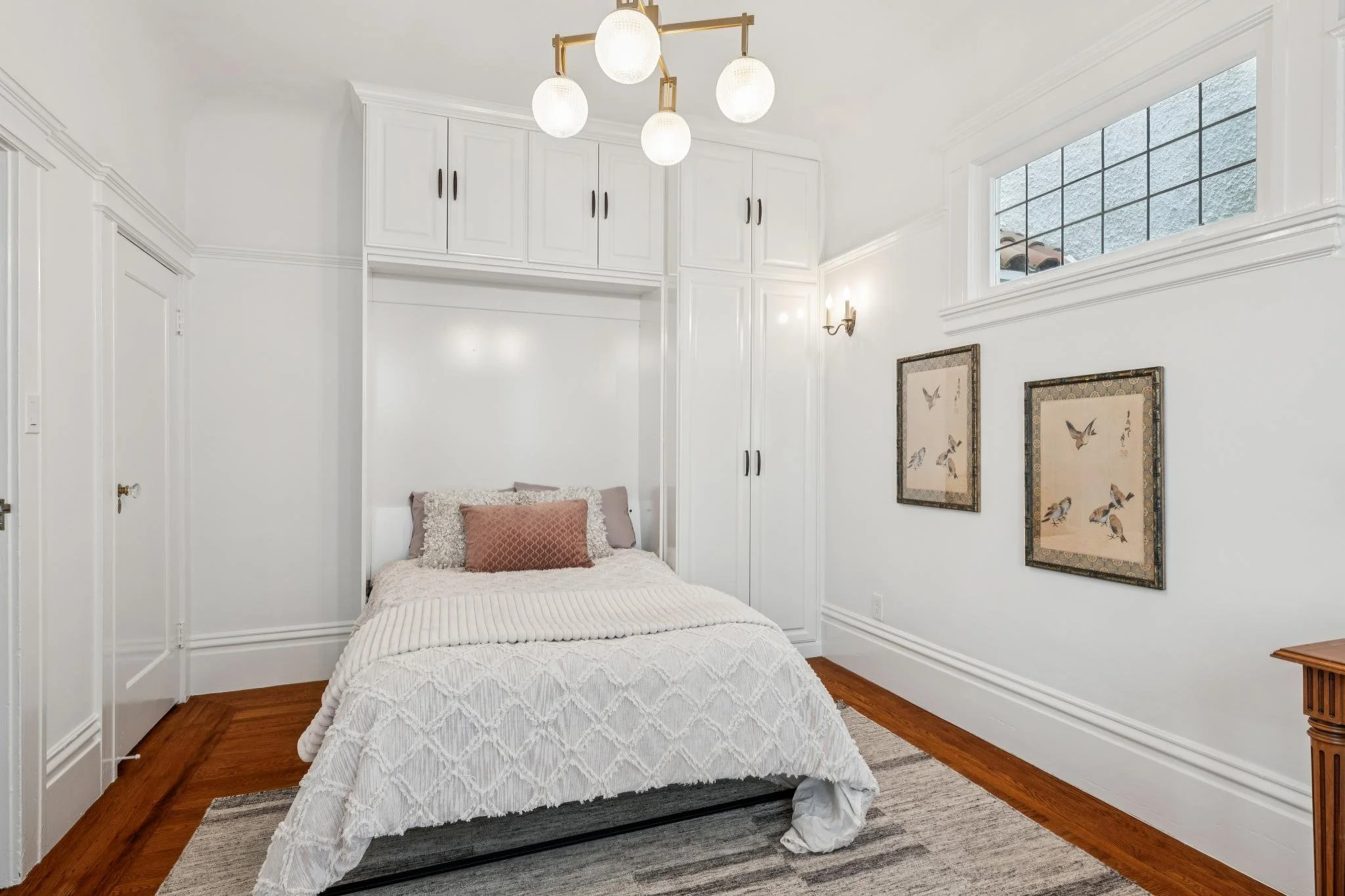 A bedroom with white walls, a bed with white bedding and pink pillows, built-in white cabinets above the bed, a window with a grid pattern, two framed bird prints on the wall, a chandelier with five white globes, a wall sconce light, and hardwood flo