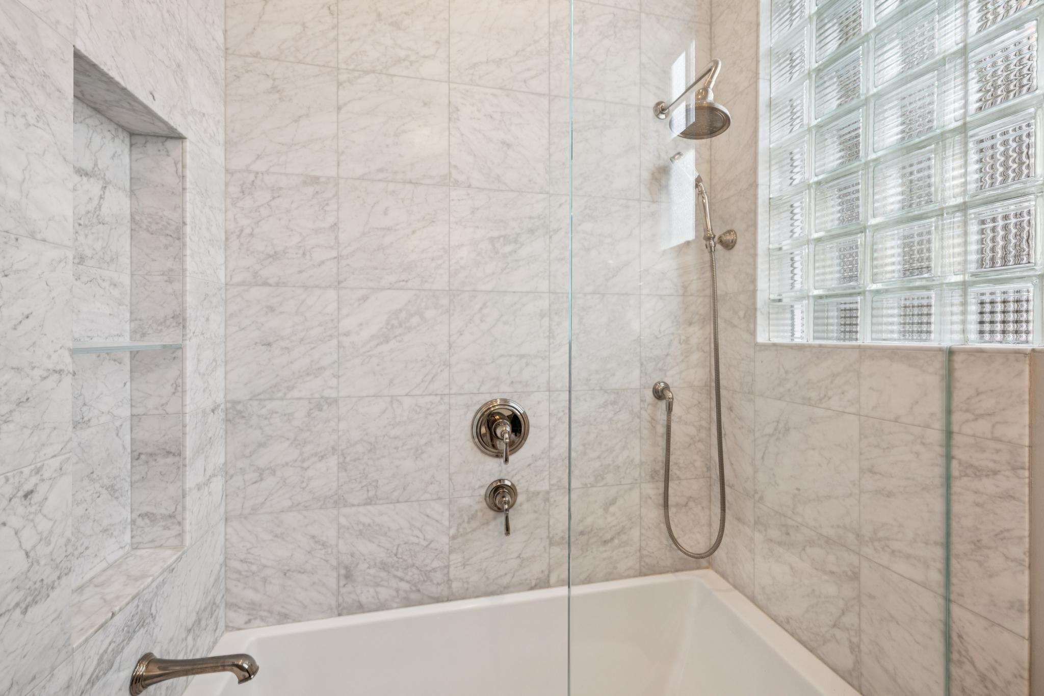 A marble shower with a built-in shelf, a glass block wall, a rainfall showerhead, a hand-held showerhead, and three control knobs.