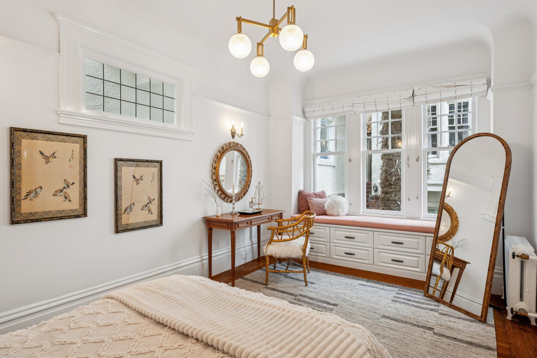A cozy reading nook with large windows, a built-in bench with cushions, and a window seat with drawers. There are vintage-style chairs, a mirror, paintings of birds, and a modern ceiling light fixture in a well-lit, white-walled room.