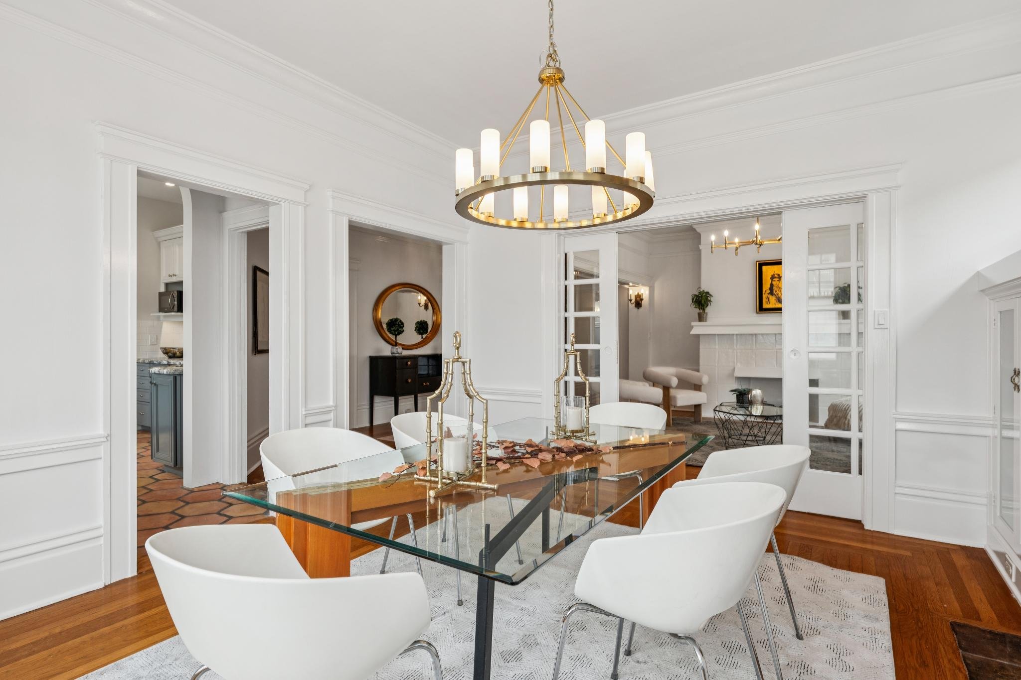 A modern dining room with a glass-top table, white chairs, and an elegant brass chandelier. The room features white walls, crown molding, and open doorways leading to a kitchen and living area. Decor includes candles, a mirror, and houseplants.