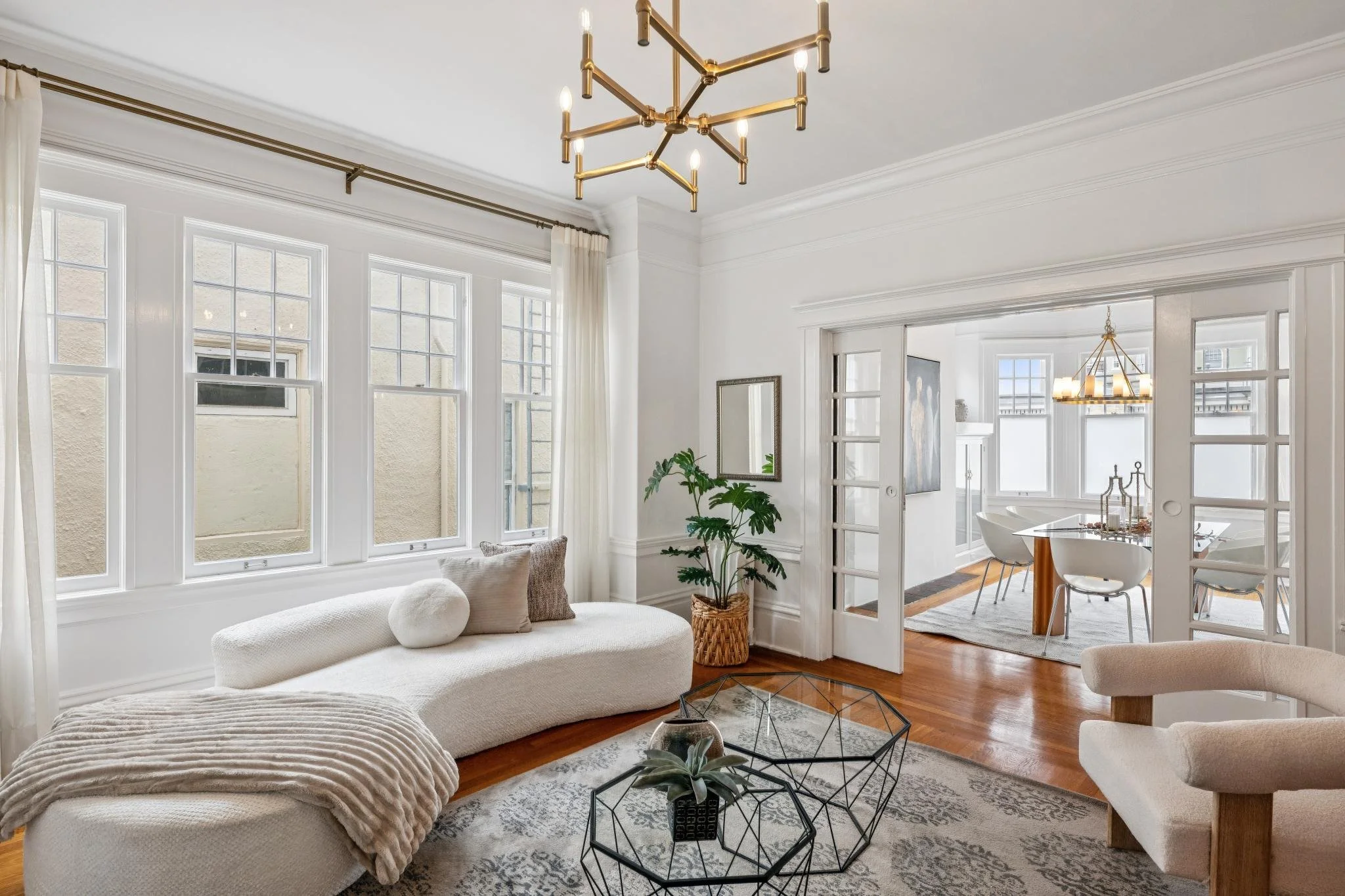 Bright living room with large windows, white walls, a white curved sofa with pillows, a glass coffee table, plants, and a dining area in the background.