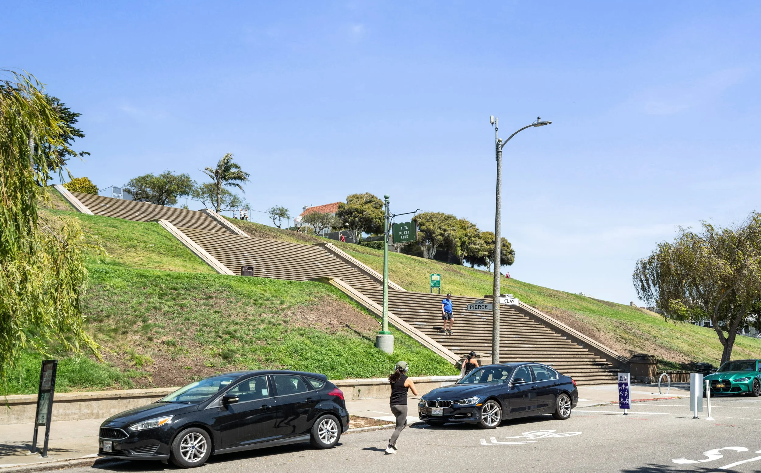 Parking lot with several parked cars, people walking, a hillside with stairs, trees, street lamps, and street signs for Alta Plaza Park and Pierce Street.