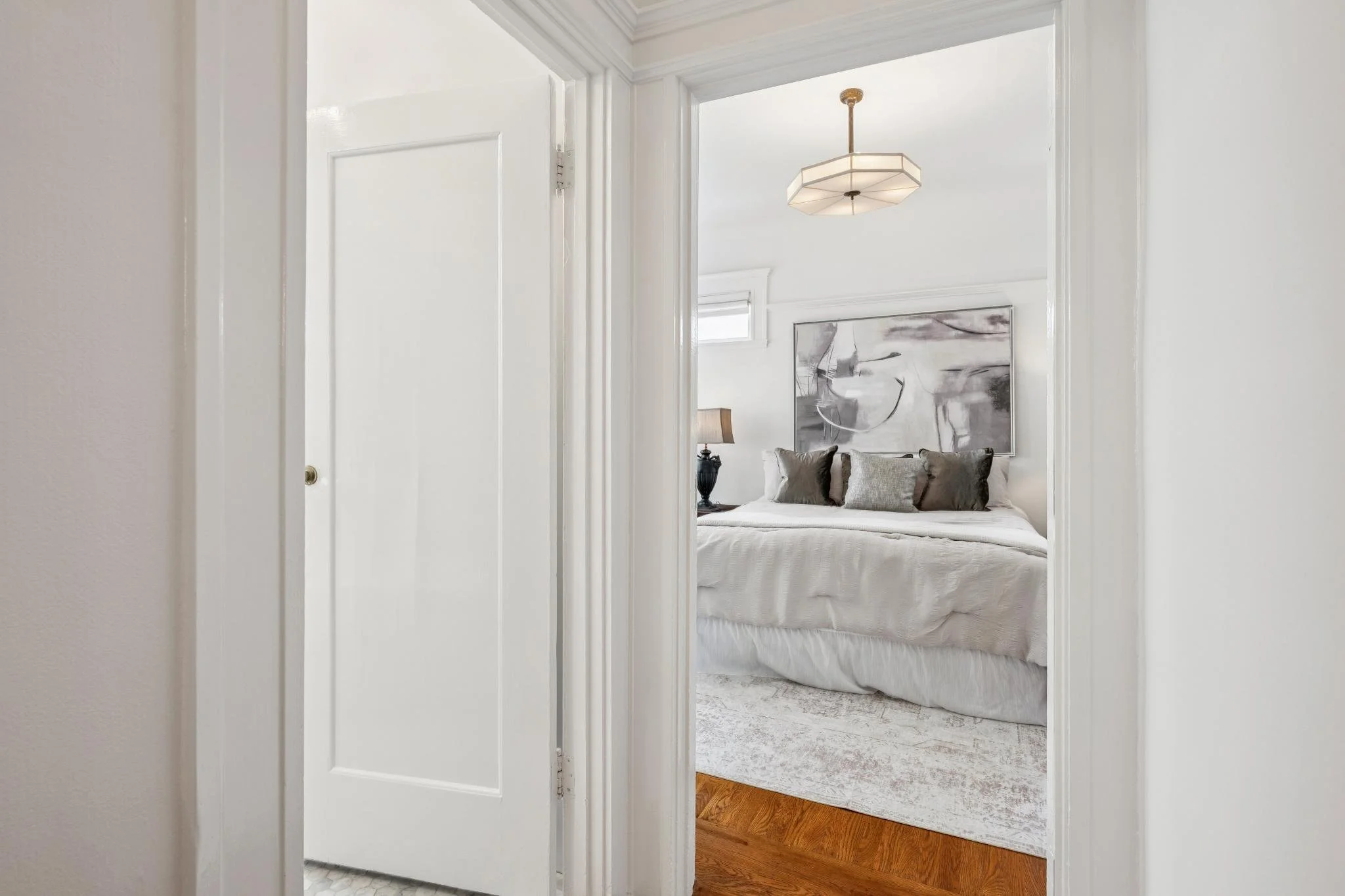 View into a bedroom from an adjoining room, showing a bed with gray pillows, artwork on the wall, a nightstand with a lamp, and wooden flooring.