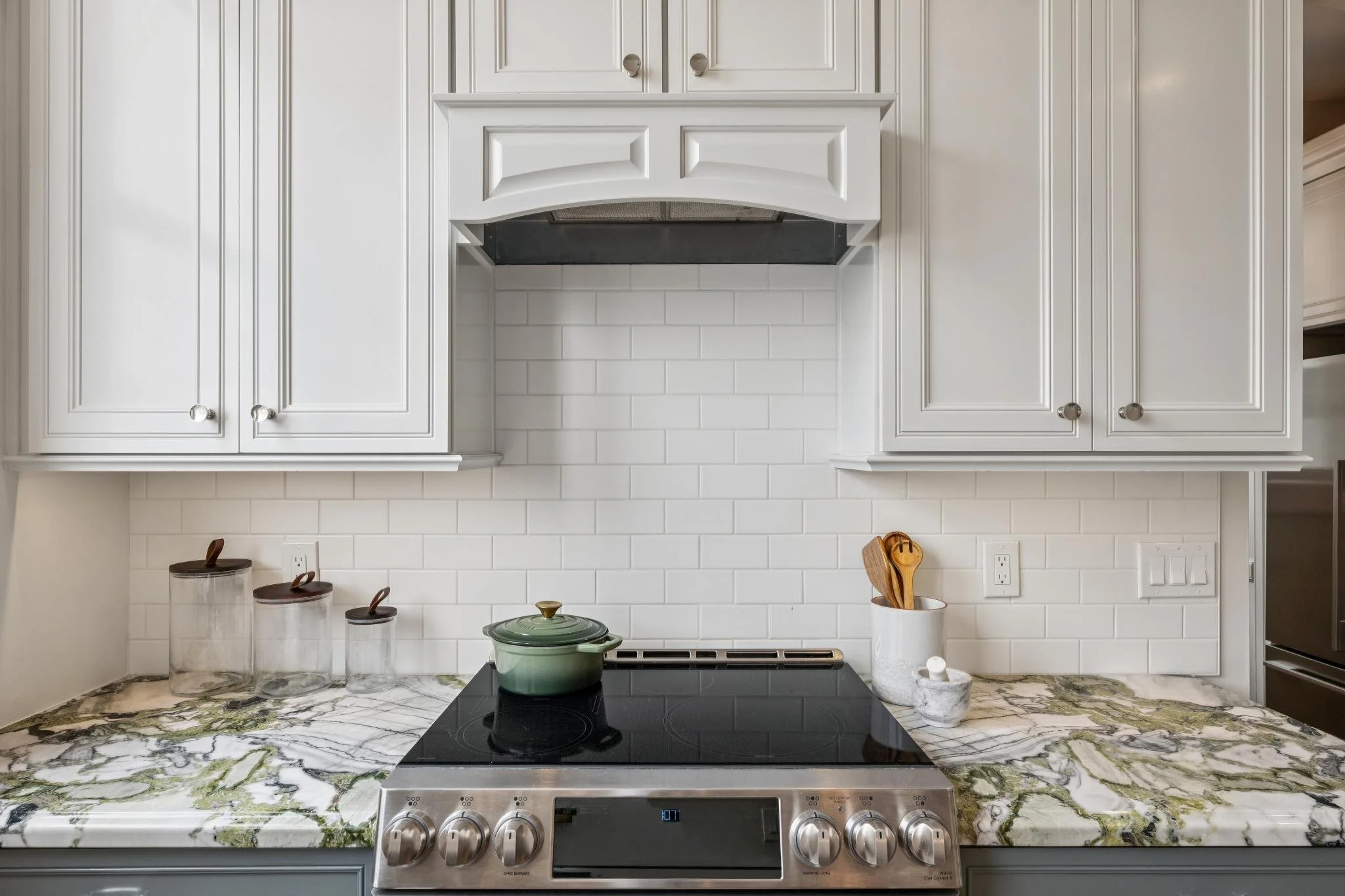 Kitchen with white cabinets, a marble countertop, a black stove with silver knobs, a green pot, and various jars with wooden spoons.