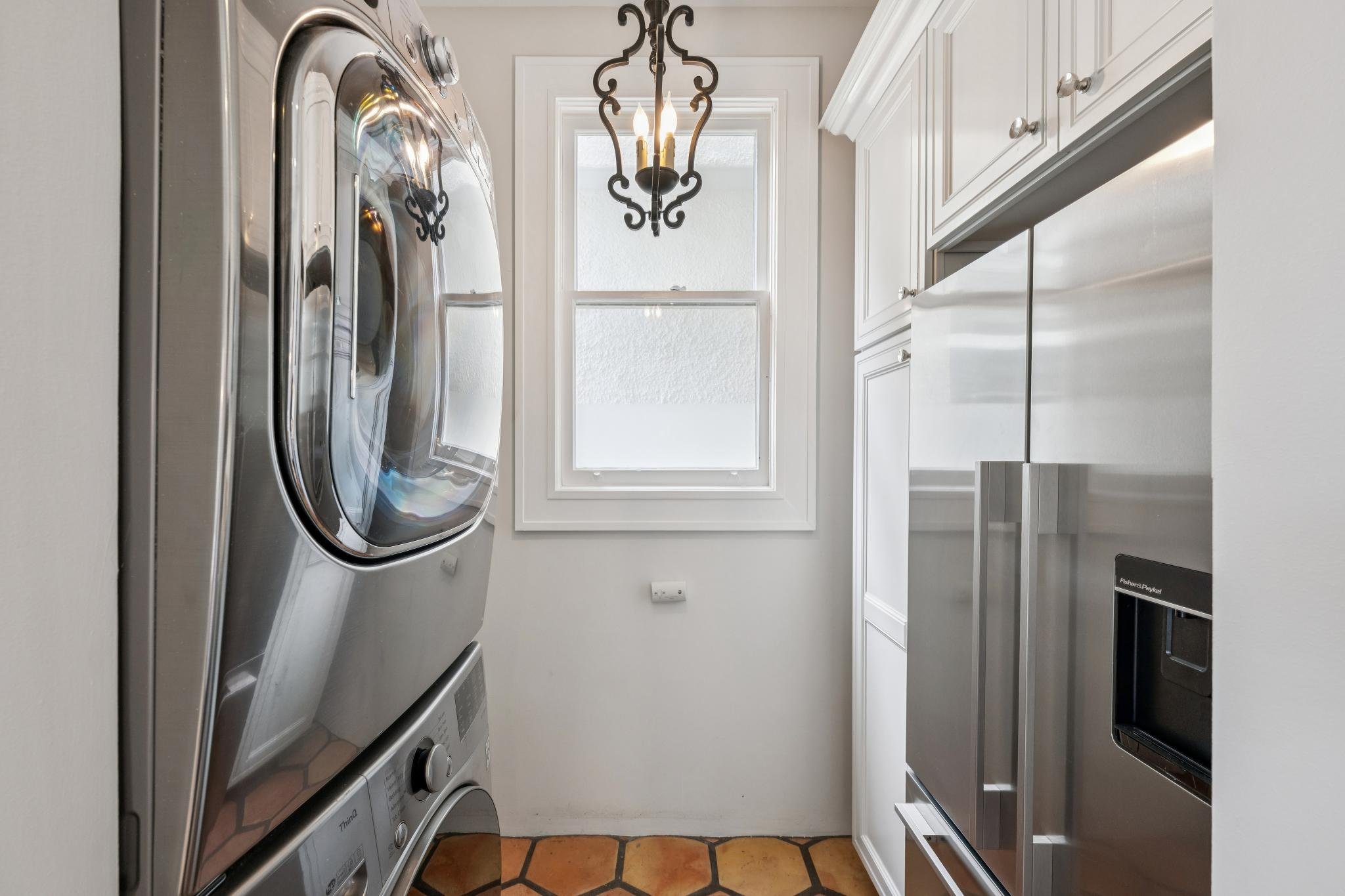 A small laundry room with a front-loading stainless steel washer and dryer on the left, white cabinets on the right, a window with frosted glass, and a black wrought iron chandelier hanging from the ceiling.