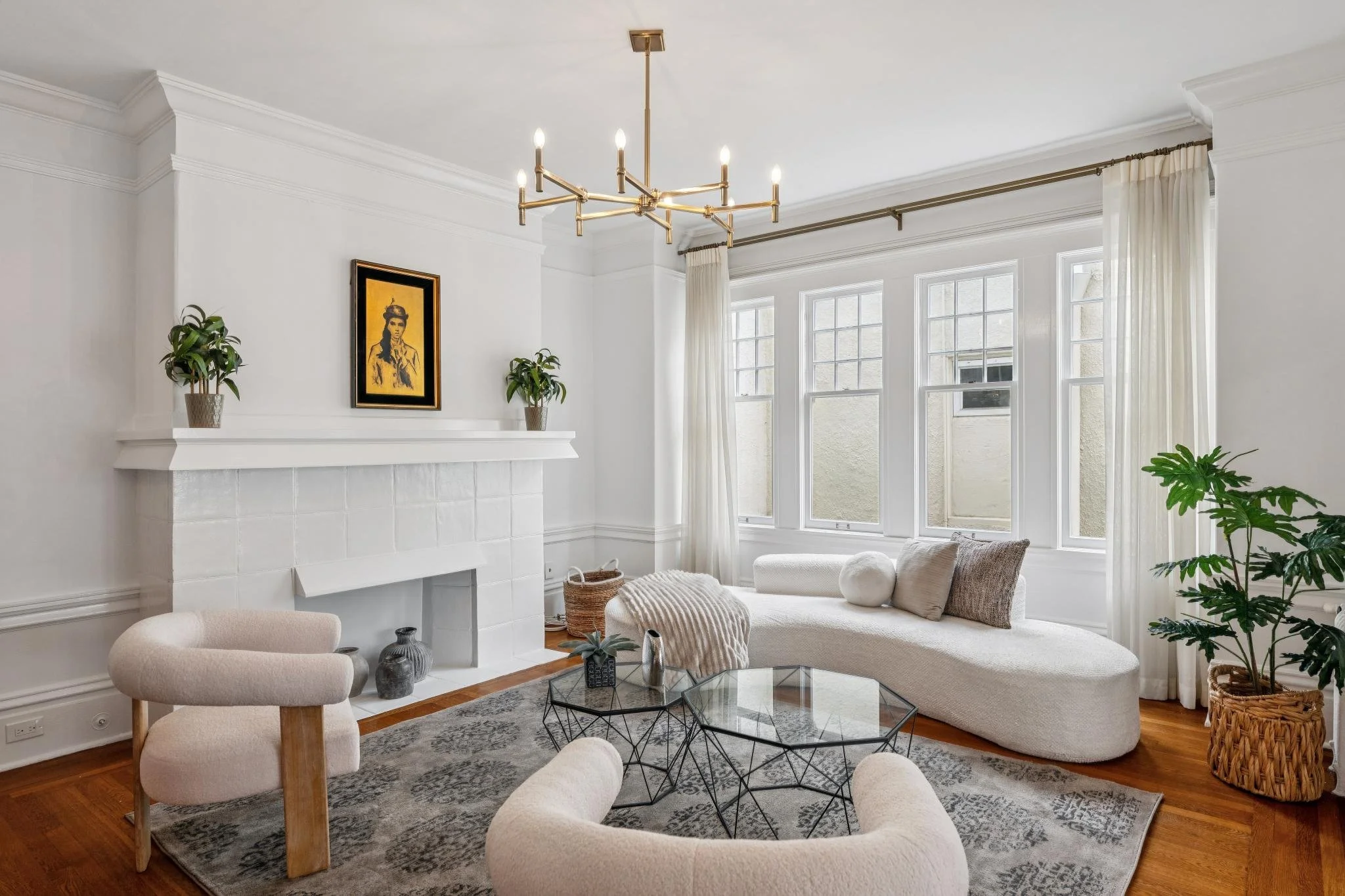 Bright living room with white walls, large windows with drapes, and modern lighting fixture. Decor includes potted plants, artwork above the fireplace, and a comfortable seating area with neutral-colored furniture and a textured rug.