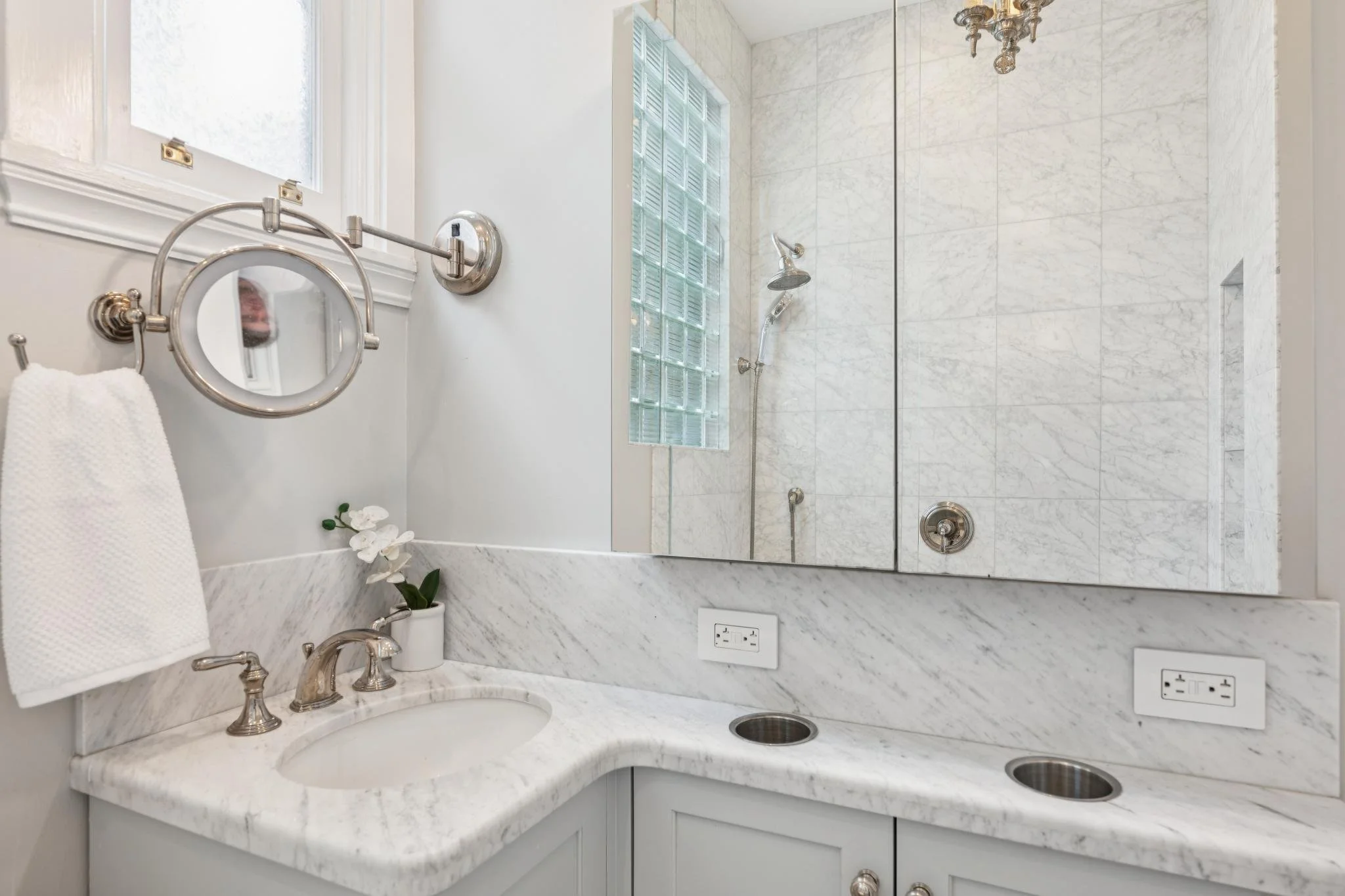 Bathroom with marble countertop and backsplash, oval sink, faucet, and mirror. A wall-mounted magnifying mirror and a towel holder with a white towel are on the wall. A potted orchid is near the sink. In the background, there is a glass-block window 
