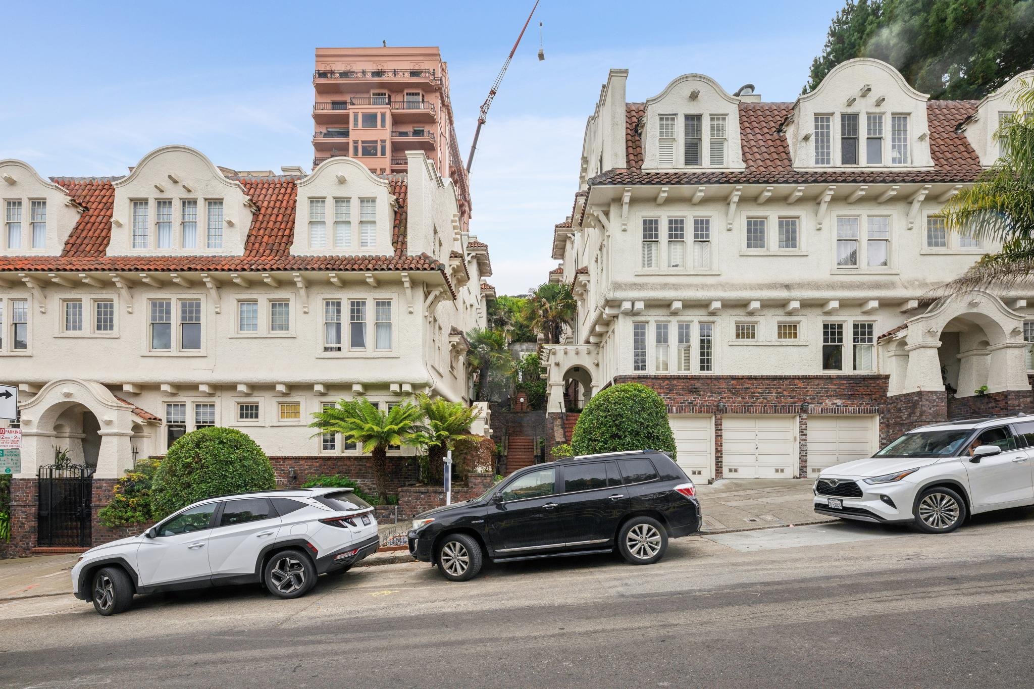 Residential street with multiple houses featuring Spanish-style architecture, white stucco walls, red tile roofs, and large windows. Several cars are parked along the street in front of the homes. A tall construction crane is visible behind the house