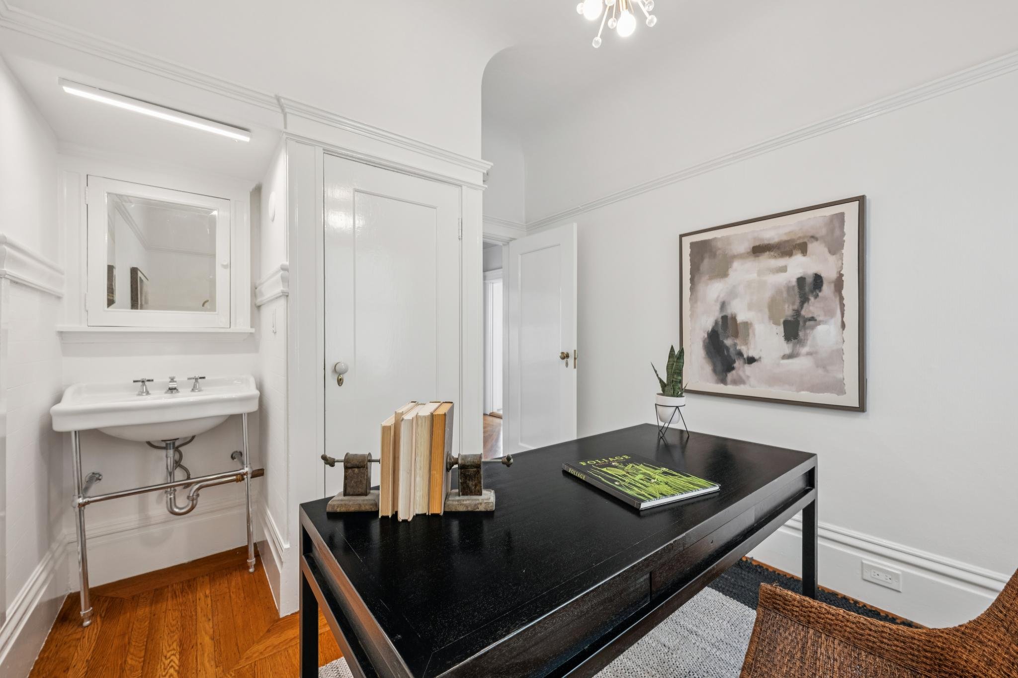Interior of a bright, minimalist room with white walls. There is a black table with a book and a plant, and a large abstract painting on the wall. A small sink is visible in the corner, and a drying rack with wooden clothespins is on the table. The c