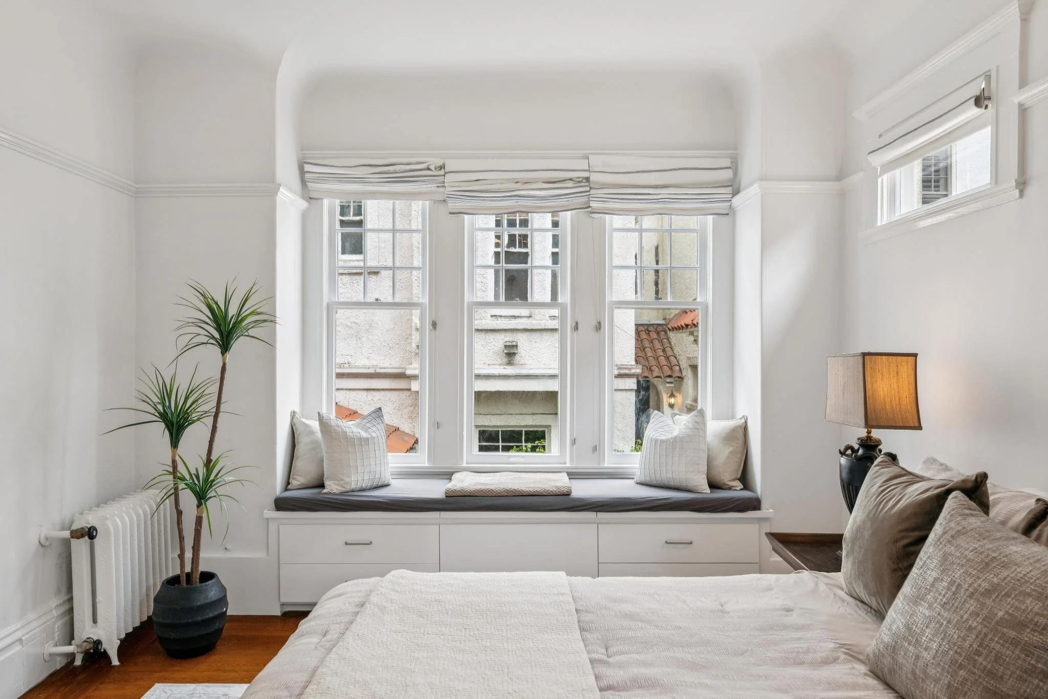 A cozy bedroom with large bay windows, decorated with striped curtains, white walls, a window seat with pillows, a tall potted plant, a radiator, and a nightstand with a table lamp.