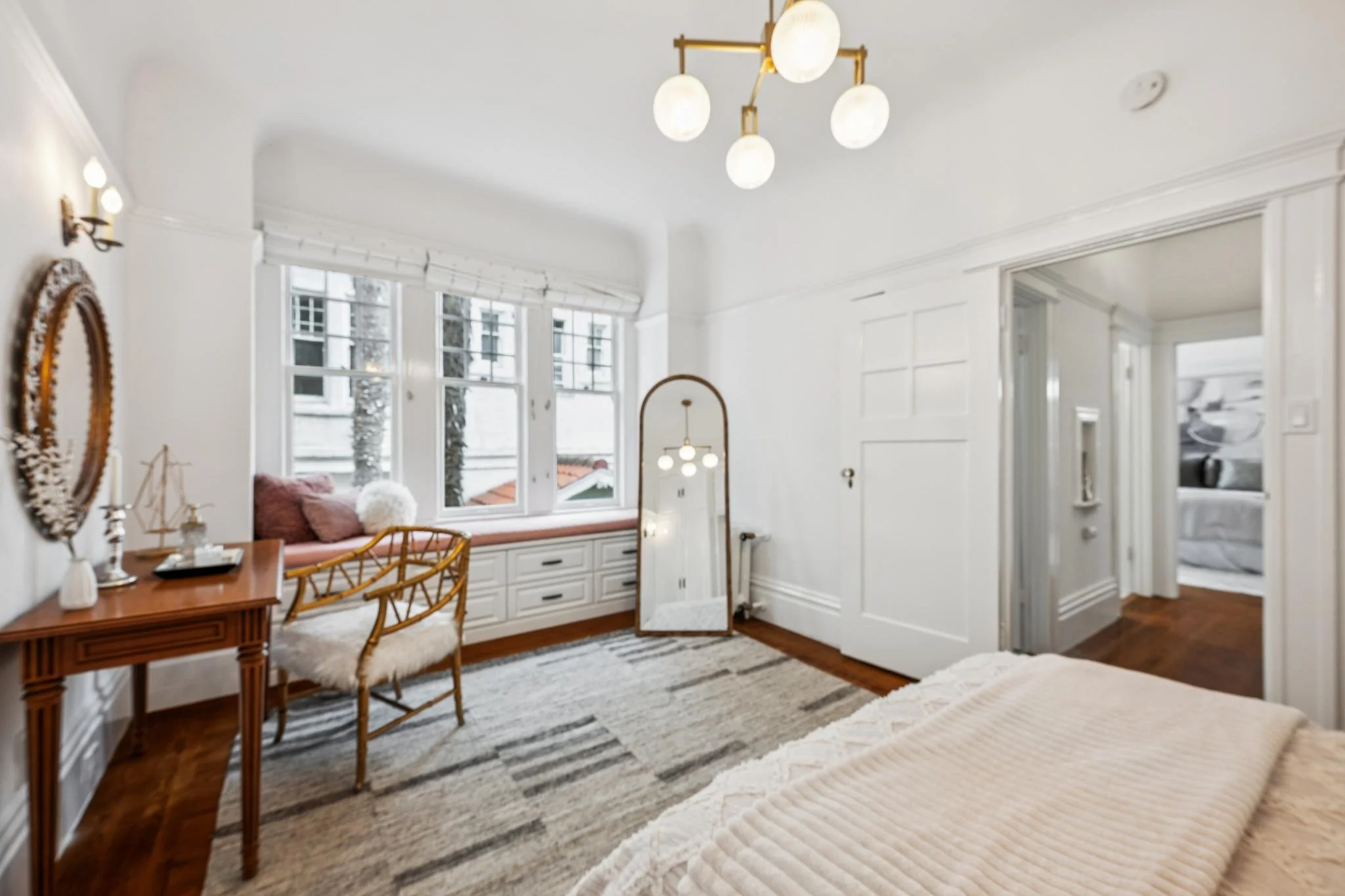 Bright living room with white walls, large windows, a wooden table with chairs, a mirror, and a bed partially visible in the foreground.