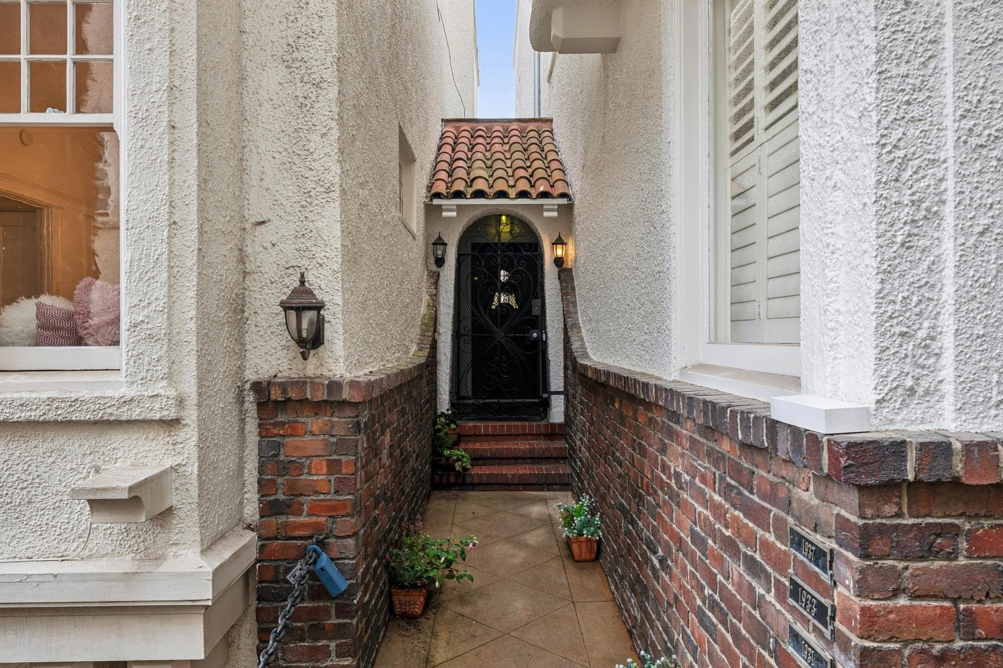 The image depicts the entrance of a house, with a black wrought iron gate set within a brick archway. To the left, there's a window with a white frame, showing pink and patterned cushions inside. The house walls are textured with a beige stucco finis