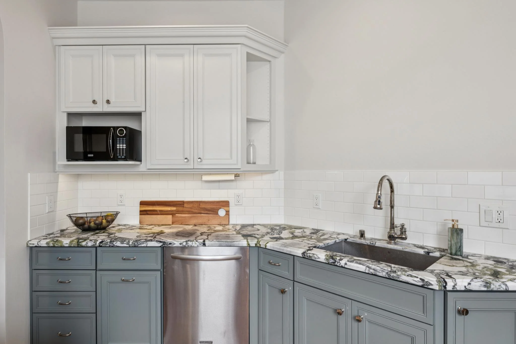 A kitchen with blue lower cabinets, white upper cabinets, a marble countertop, a black microwave, a black toaster oven, and a section of white subway tile backsplash.