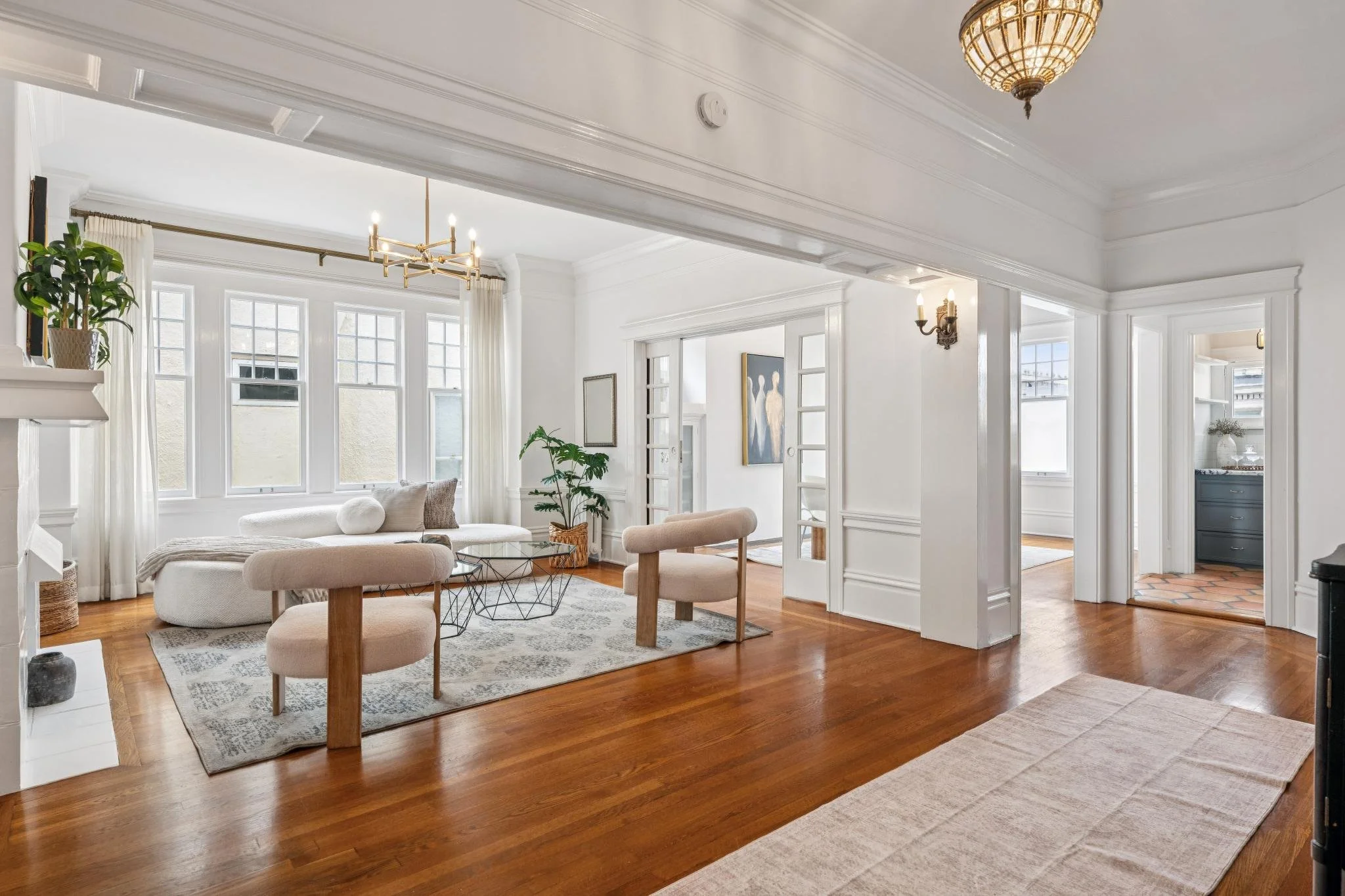 Bright, airy living room with white walls, large windows, and hardwood floors. Features a beige sofa, round coffee table, potted plants, and modern artwork. Elegant lighting fixtures include a chandelier and wall sconces.
