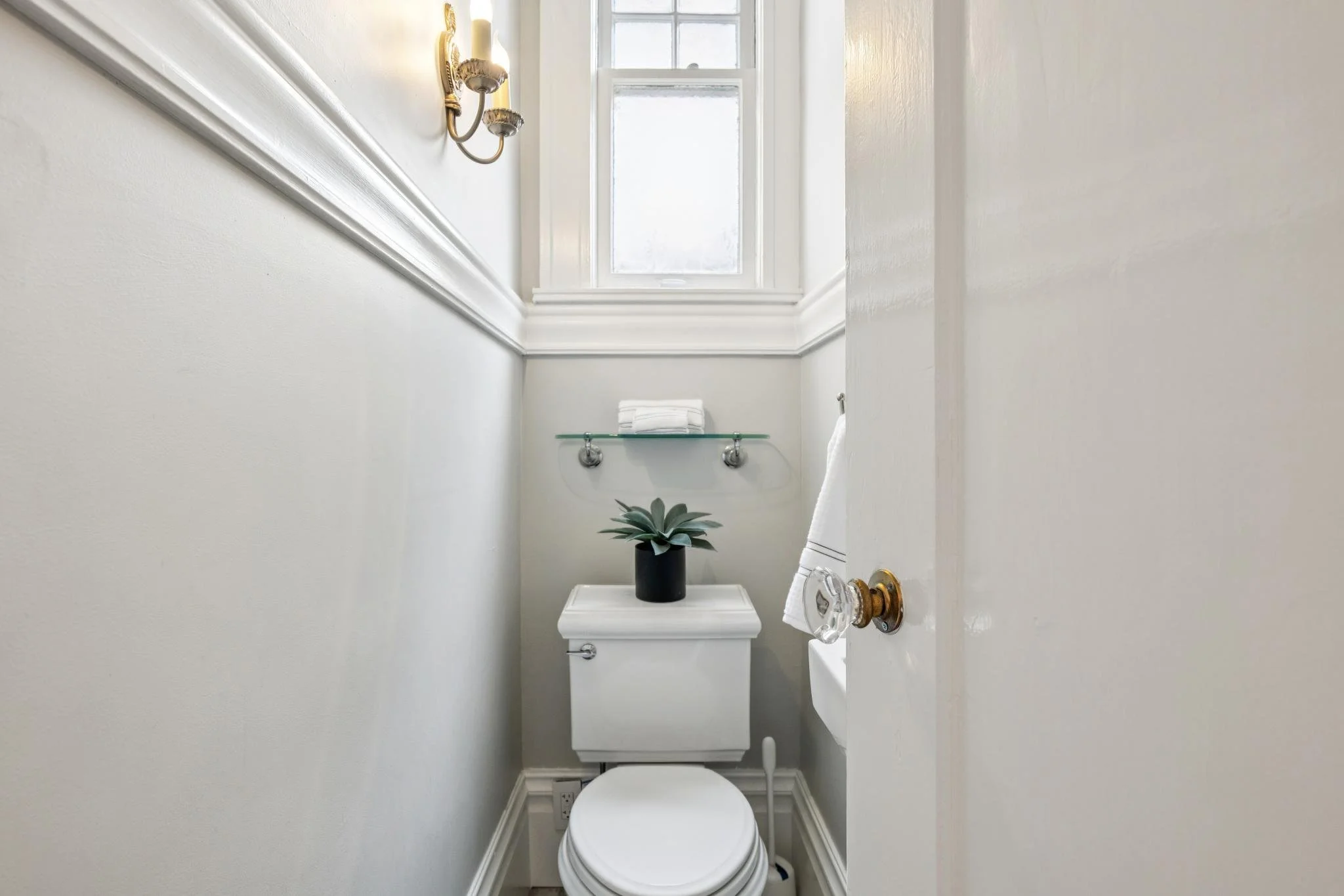 Small bathroom with a white toilet, a glass shelf with folded towels, a potted plant, a window, and wall-mounted light fixtures.
