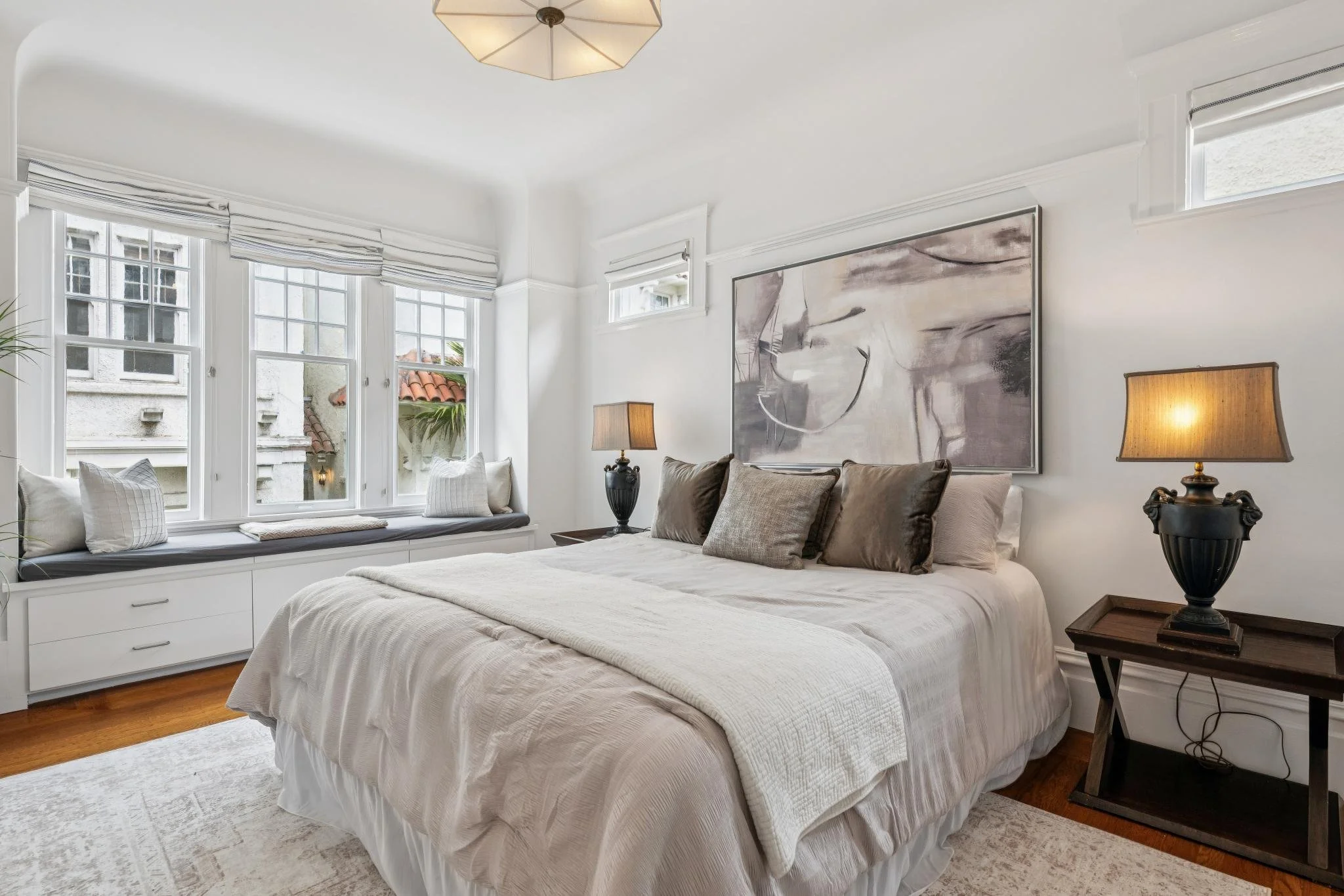 A bright bedroom with a large white bed, dark accent pillows, a wooden nightstand with a black lamp, large windows with white shutters and curtains, and artwork on the wall.