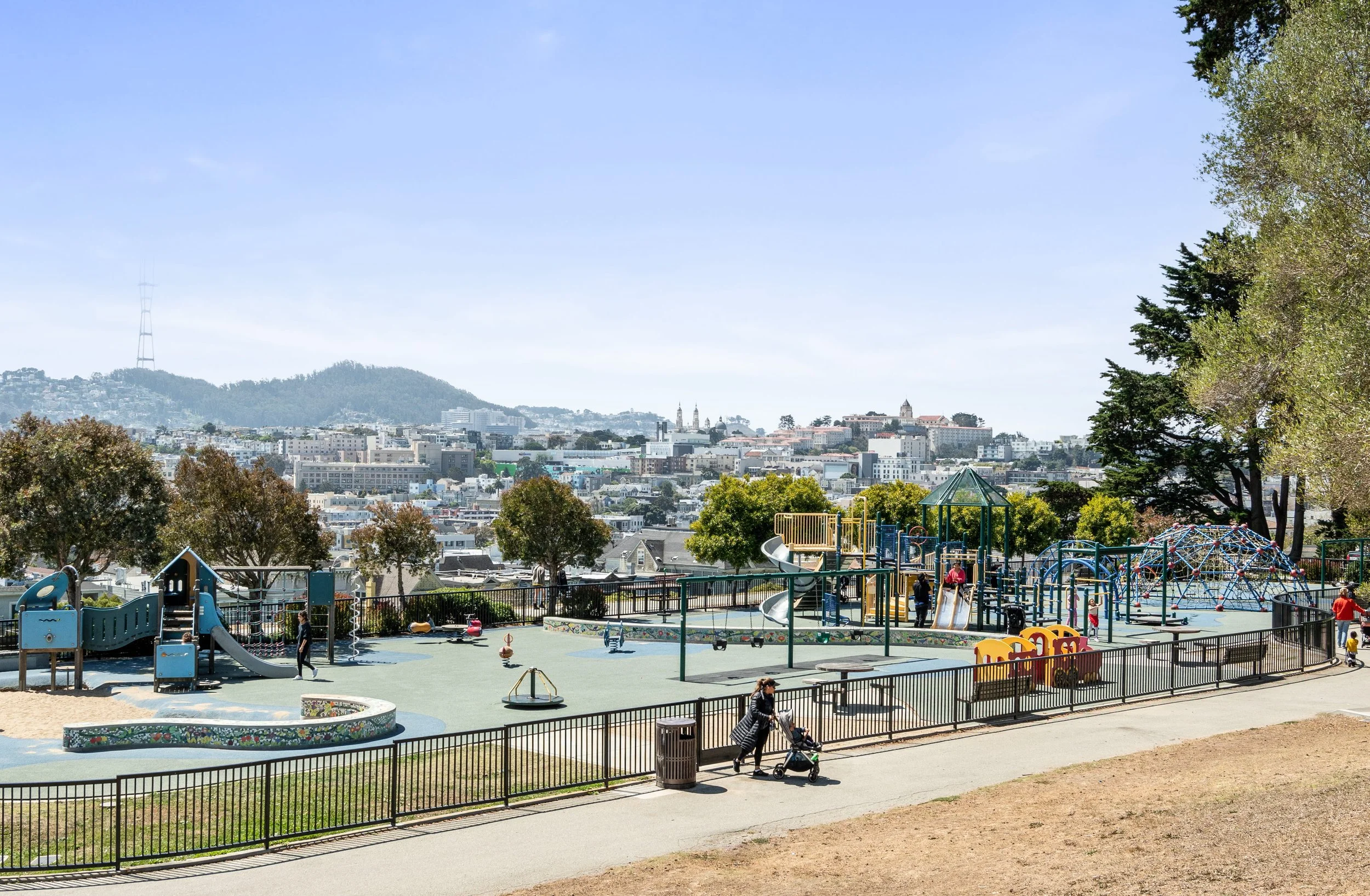 A children's playground with slides, climbing structures, and swings, surrounded by a fence, with people, trees, and a city skyline with hills and buildings in the background on a clear day.