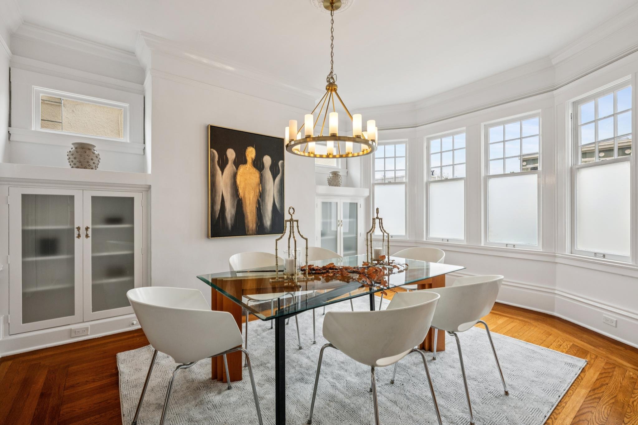 A bright dining room with large bay windows, white walls, and a wooden floor. There is a glass-top dining table with a wooden base surrounded by white chairs. A modern chandelier hangs from the ceiling, and artwork on the wall depicts abstract figures. There's a white cabinet with glass doors and decorative vases.