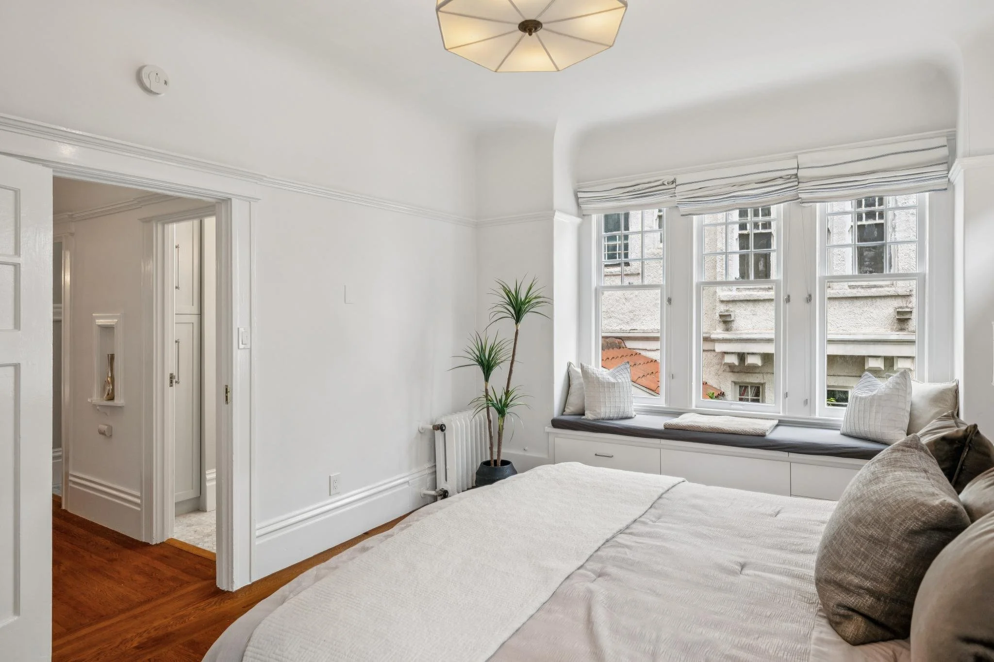 A bright bedroom with a large window seat adorned with cushions and pillows, white walls, hardwood floors, and a ceiling light. There is a tall plant near the window and a glimpse of a room with white decor in an adjacent space.