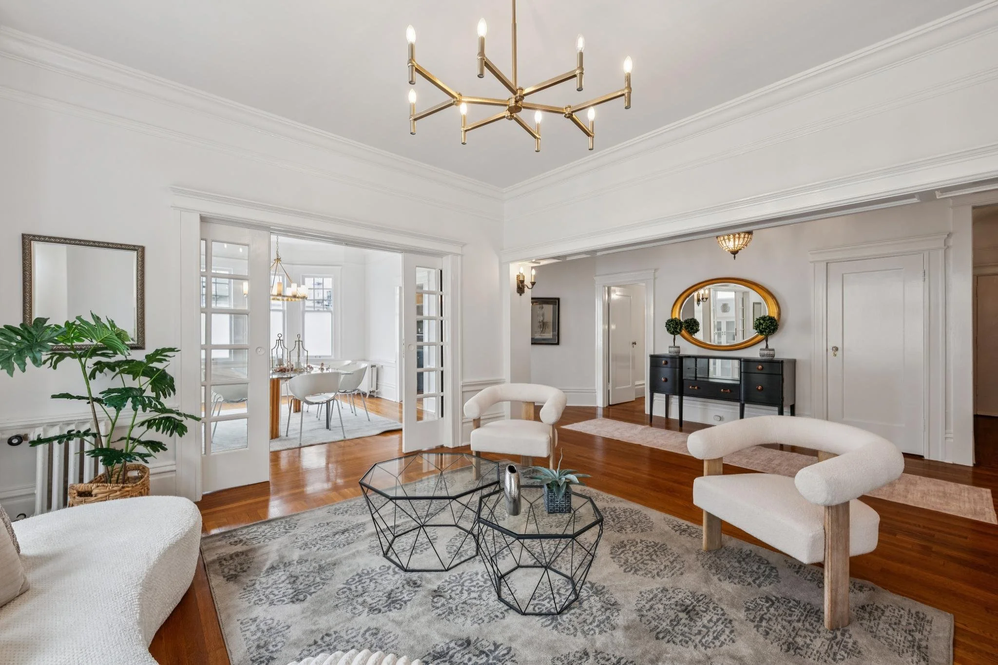 Living room with white walls, hardwood floors, elegant chandelier, mirror, black console table with two small topiary plants, white armchairs, glass coffee table, large houseplant, and a dining area visible through double glass doors.