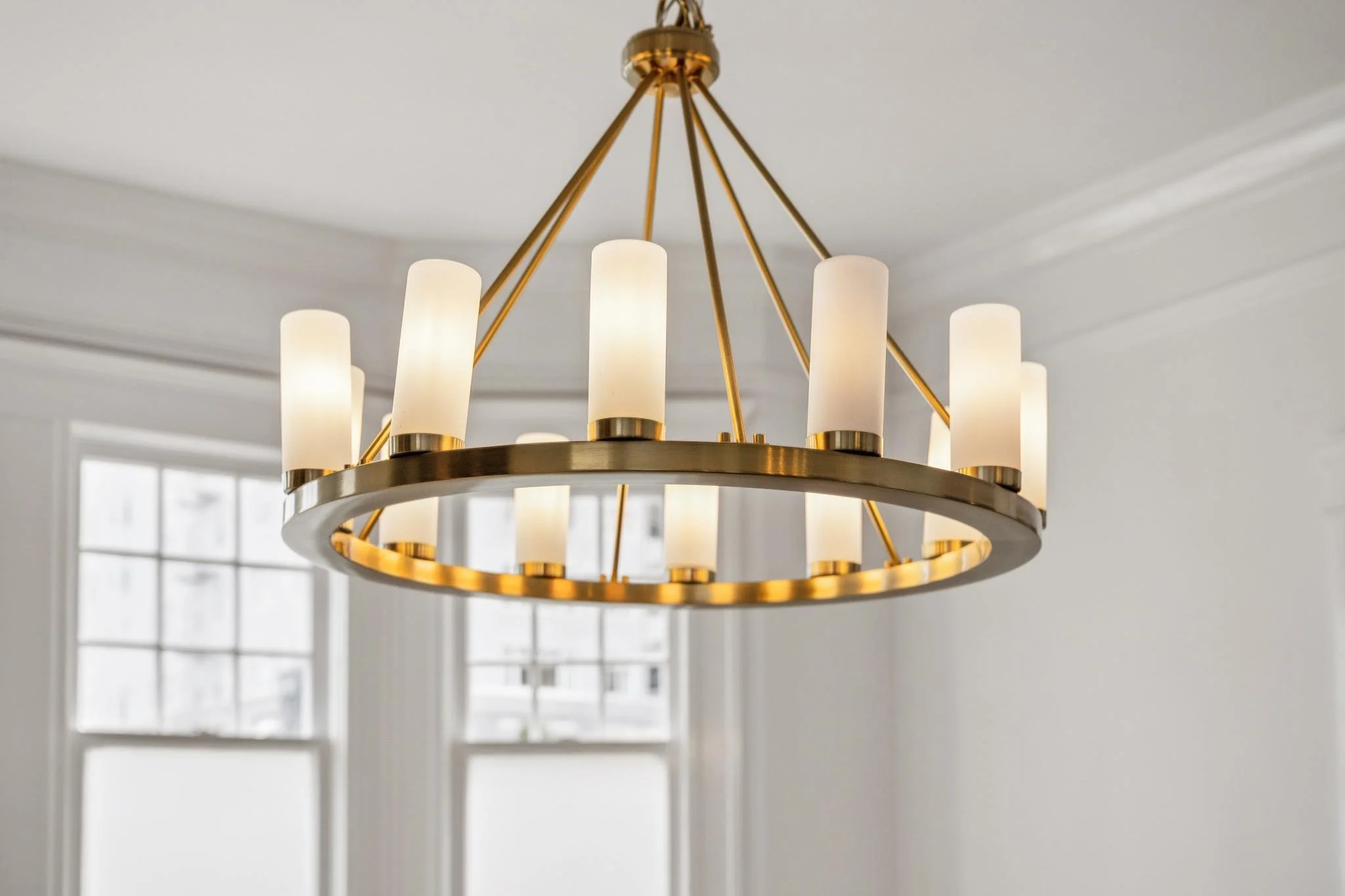 A modern chandelier with a circular gold frame and white cylindrical lampshades hanging from a white ceiling in a bright room with large windows.