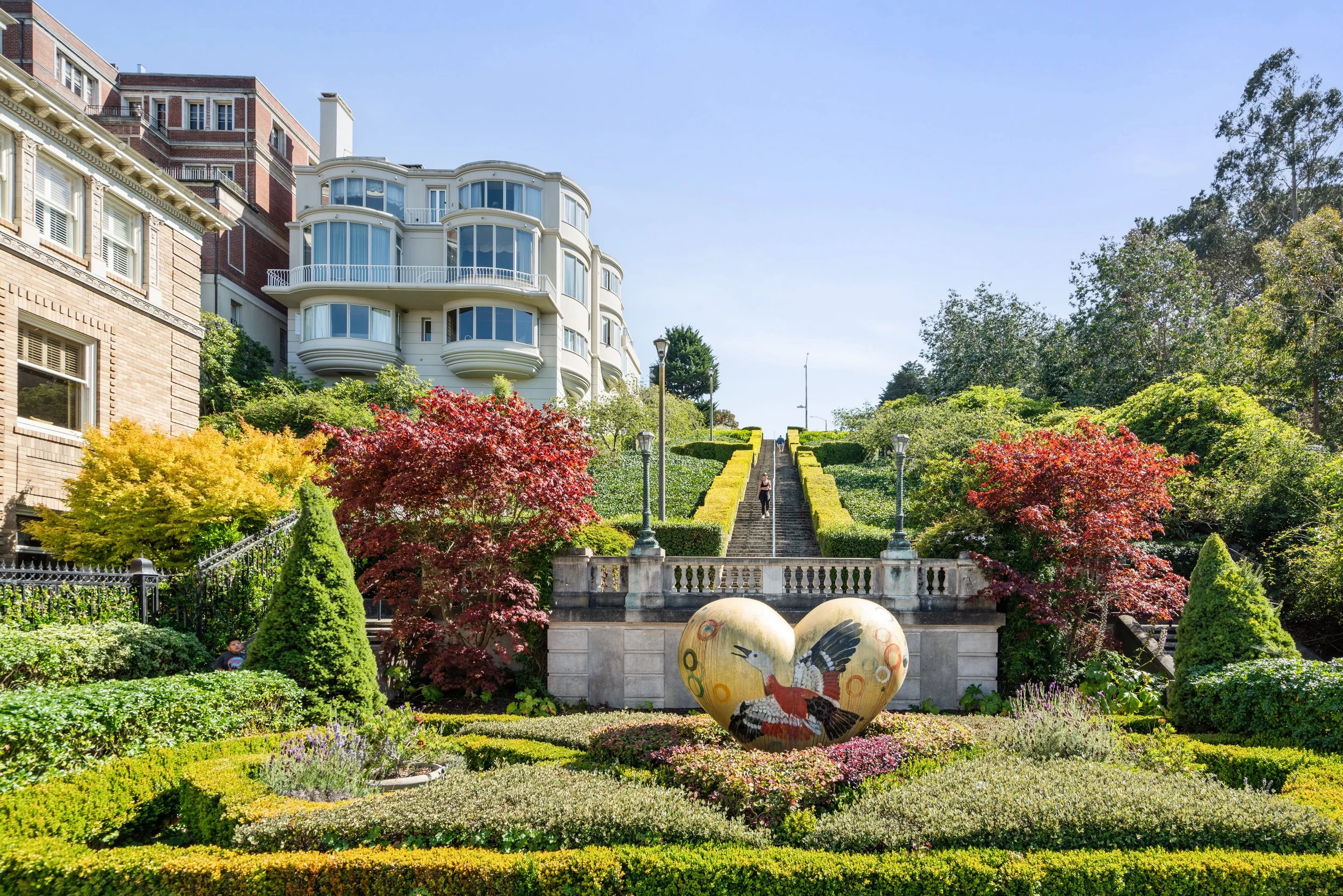 A beautifully maintained urban park featuring vibrant fall foliage, decorative ikebana-style flower beds, modern sculptures, and steps leading to residential buildings on a sunny day.