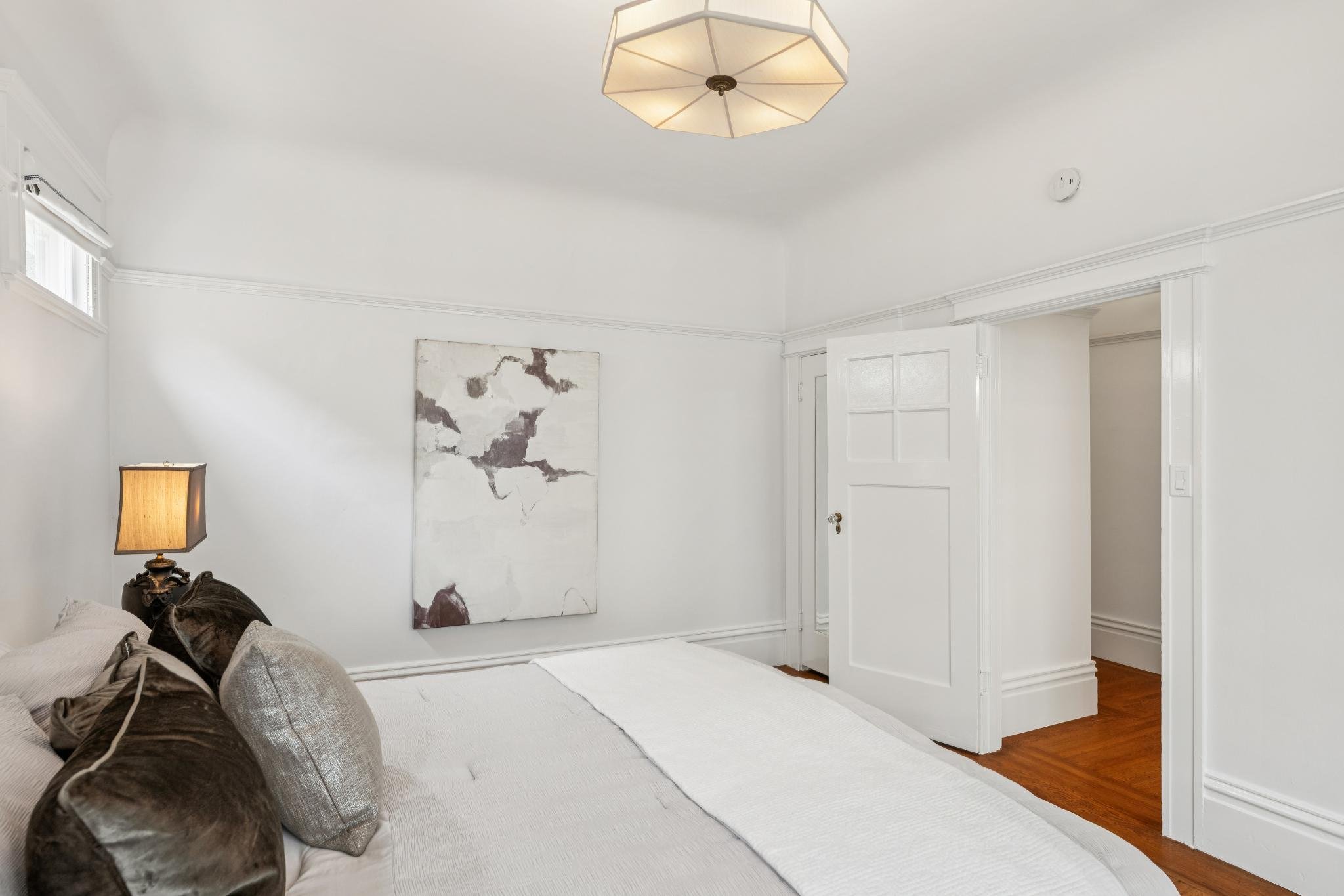 A bedroom with a white bed, decorative pillows, a table lamp, a wall art piece, and a door. The room has white walls with crown molding and wooden flooring.